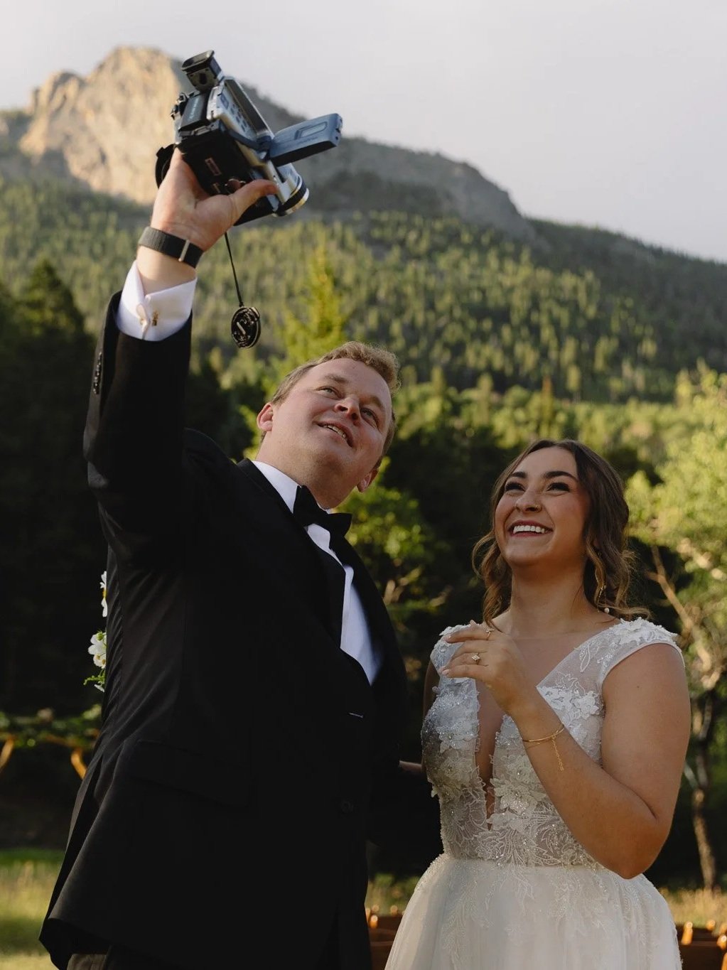 A newlywed couple taking a selfie outdoors with a mountain and greenery in the background, the groom holding a camera and both smiling. Estes Park, Colorado Wedding Photographer.