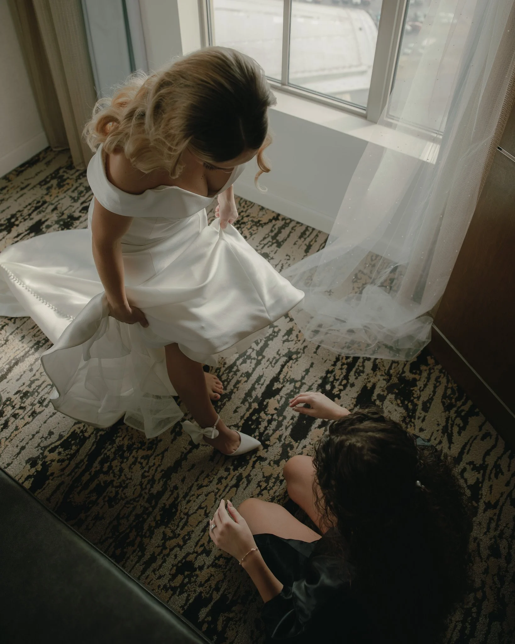 A woman in a white wedding dress is trying on white high-heeled shoes while another woman, dressed in black, sits on the floor nearby, adjusting her shoe near a window with sheer curtains.