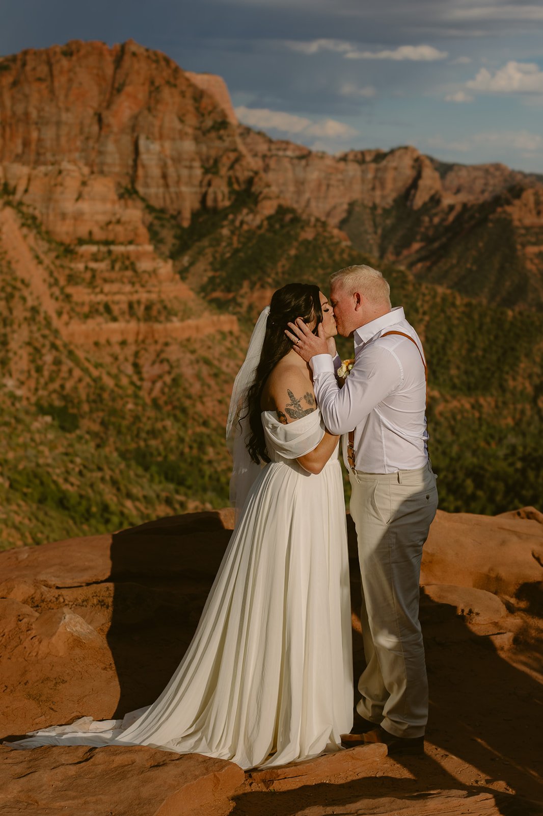 A bride and groom in wedding attire sharing a kiss on a rocky ledge with a mountainous desert landscape in the background.