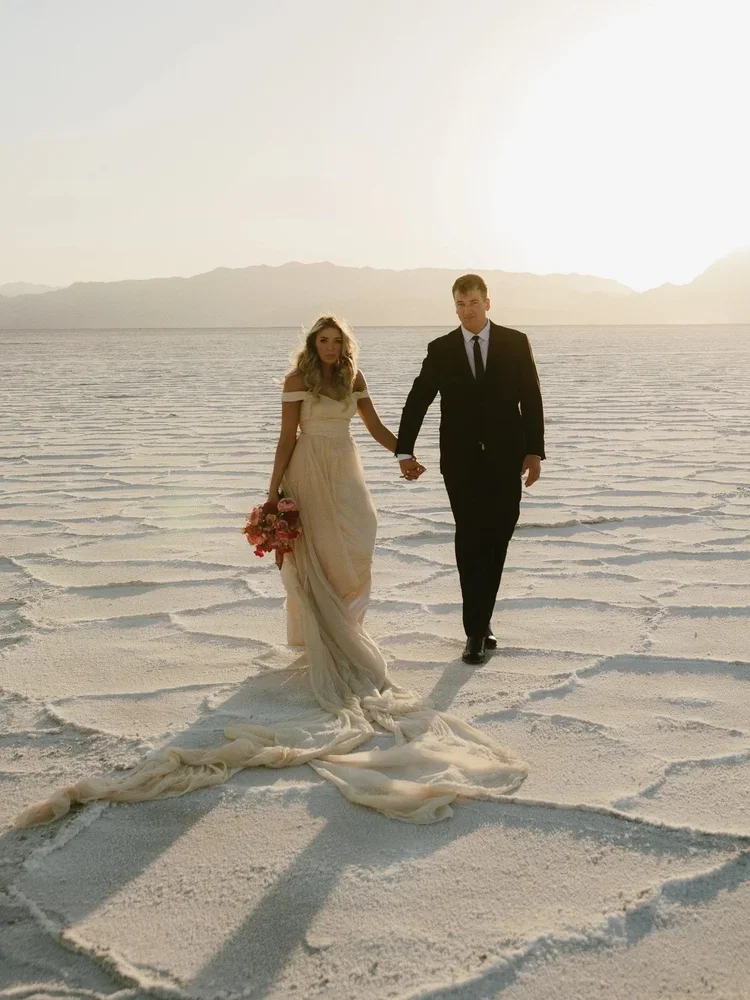 A bride and groom walking hand-in-hand across a salt desert at sunset, with mountains in the background.