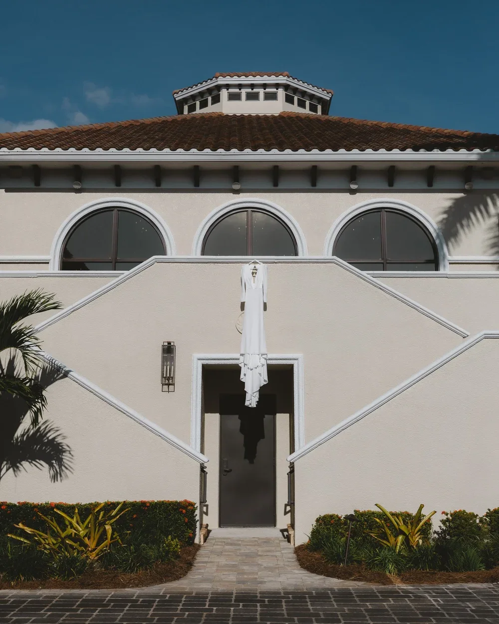 Exterior of a cream-colored building with arched windows, a staircase leading to a door, and a white dress hanging in the center, with landscaped bushes and a cloudy blue sky.