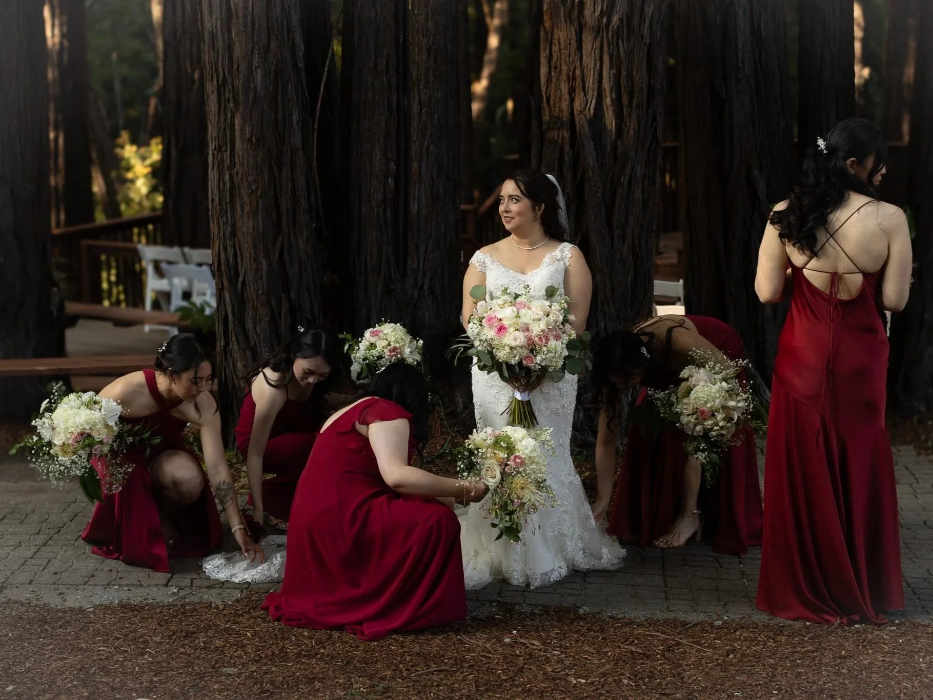 A bride in a white wedding dress holding a bouquet of flowers standing amidst bridesmaids in red dresses arranging flowers in a forest setting.