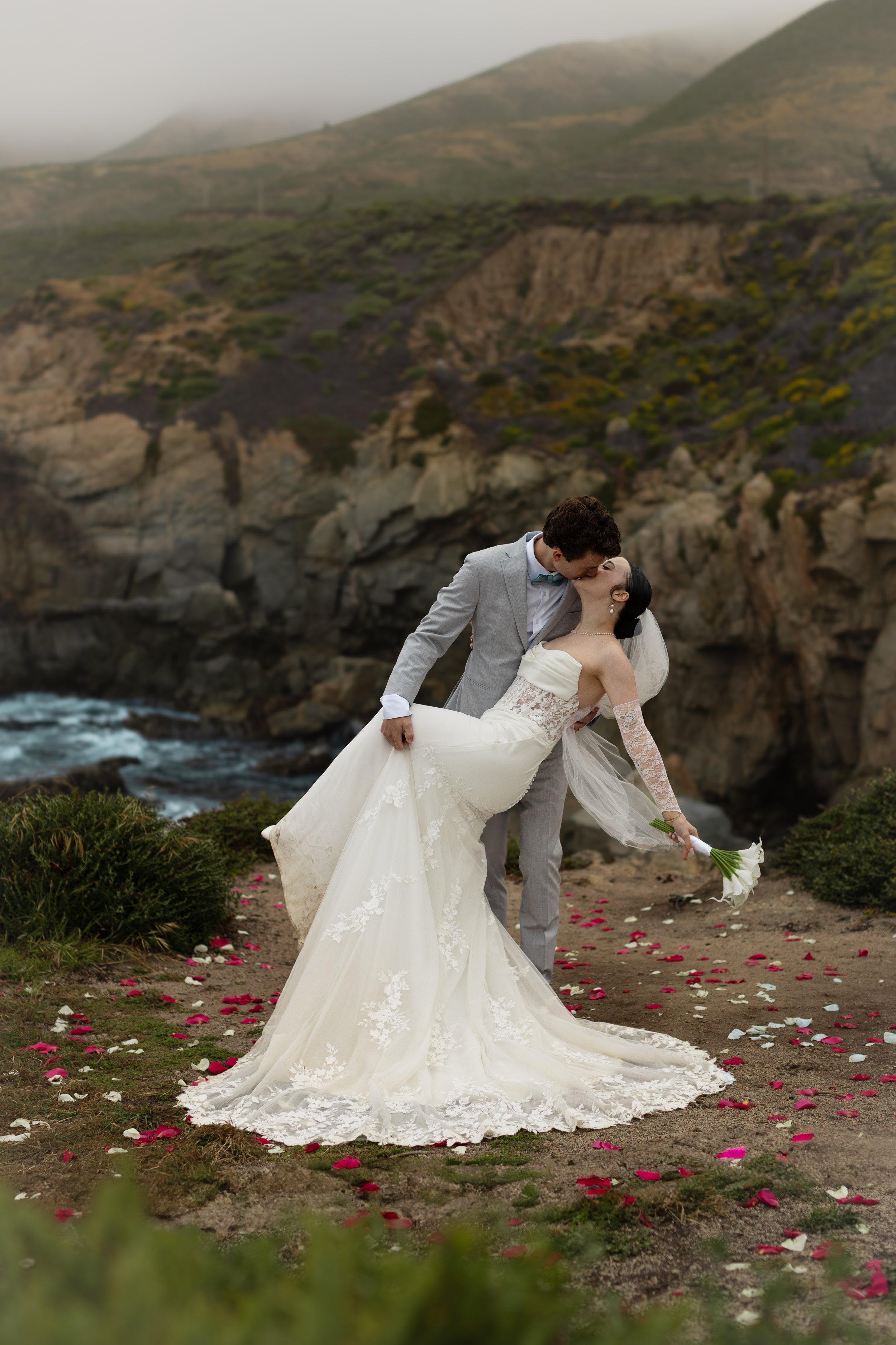 A bride and groom sharing a kiss outdoors on a rocky cliffside with a waterfall and hills in the background; the bride is wearing a white wedding gown and veil, and the groom is in a gray suit. Rose petals are scattered on the ground.