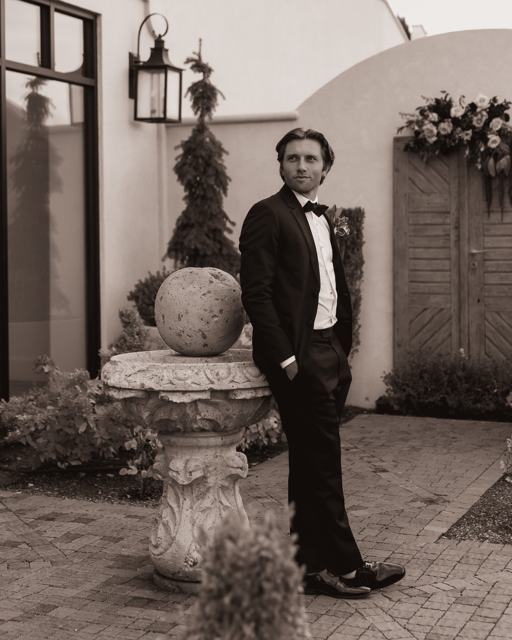 A man in a tuxedo leaning against an ornate stone pedestal with a round stone ball on top, outdoors in a garden or courtyard setting.