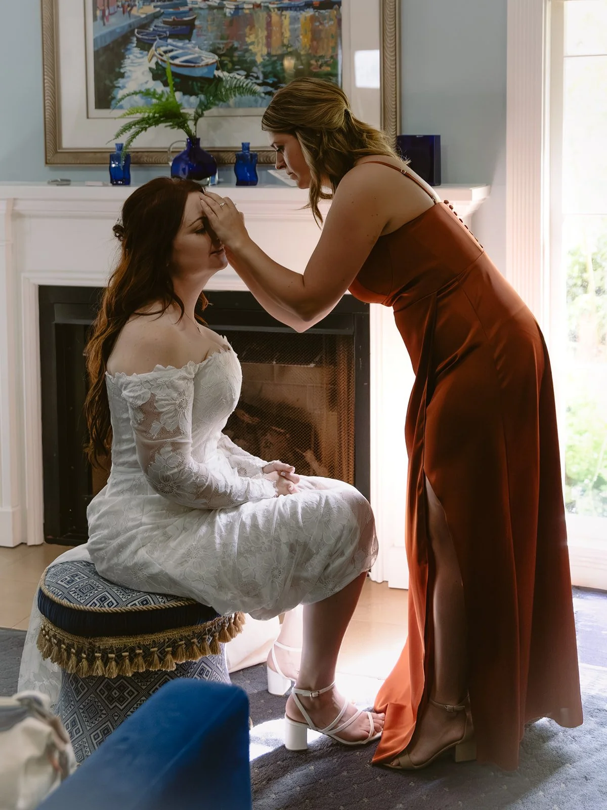 A woman in a rust-colored dress gently touches a bride's forehead in a moment of emotional support inside a bright, decorated room with a fireplace, paintings, and large windows.