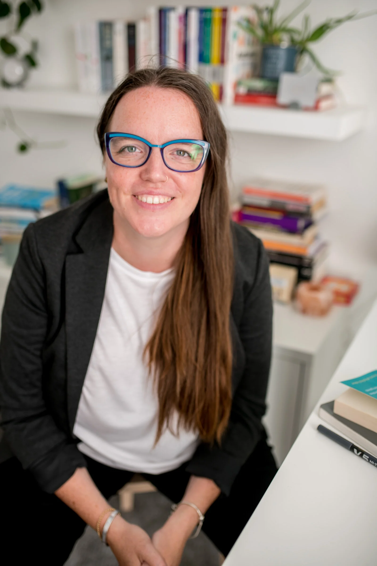 A smiling woman with long brown hair and glasses sitting at a desk in an office or study space, with bookshelves and potted plants in the background.