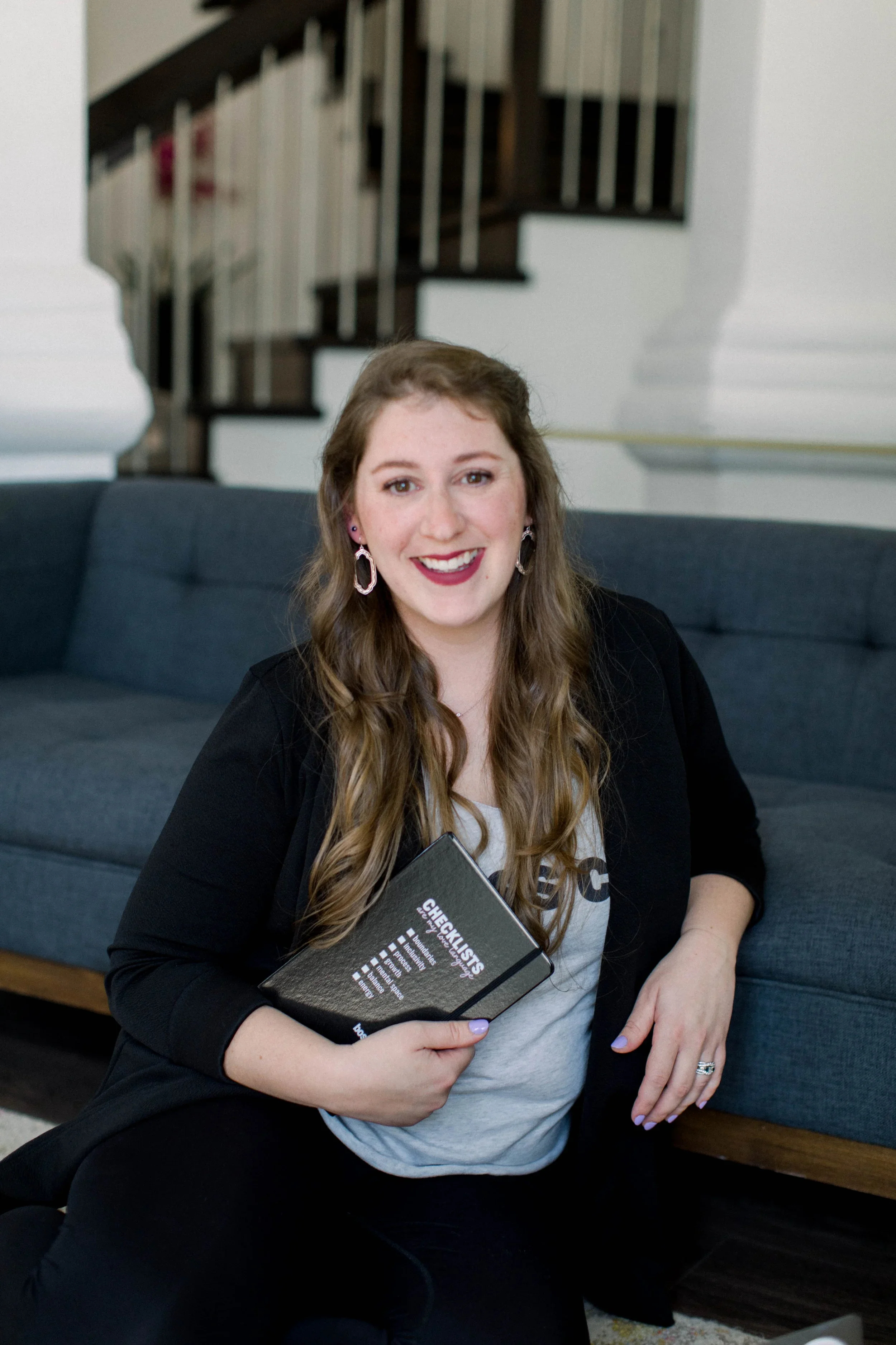 A smiling young woman with long wavy hair, wearing earrings, a black blazer, and a light-colored shirt, holding a black checklists notebook, sitting on the floor next to a dark gray couch in a home interior.