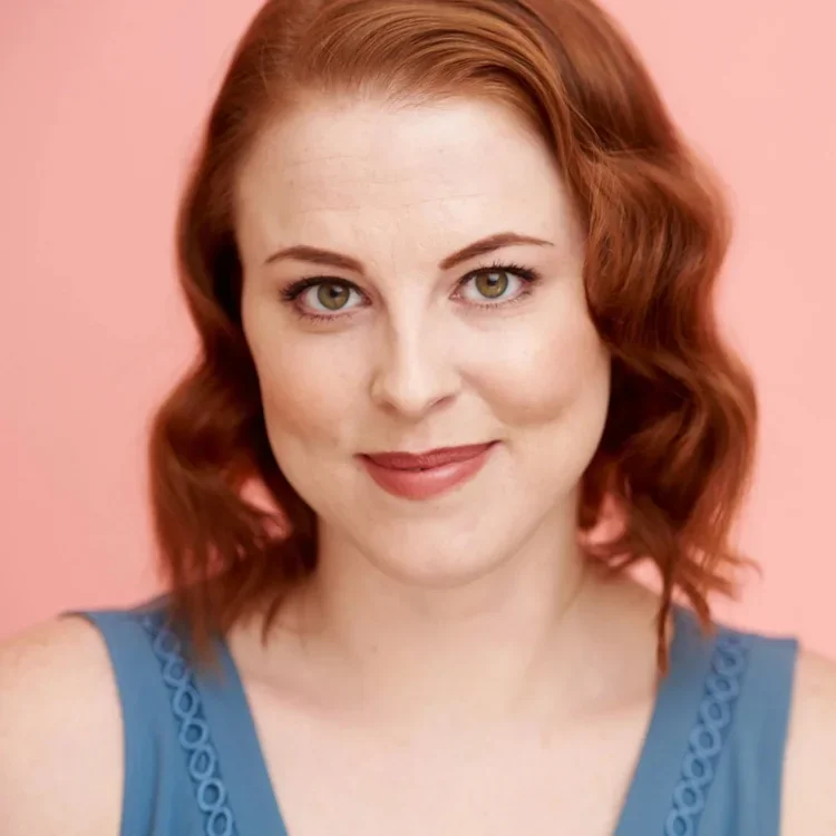 A young woman with curly red hair and green eyes, wearing a sleeveless blue top, smiling against a pink background.
