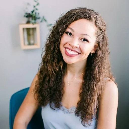 A young woman with curly brown hair and a big smile, wearing a light blue top, sitting on a blue chair in a room with a light gray wall and a small bookshelf with a plant in the background.