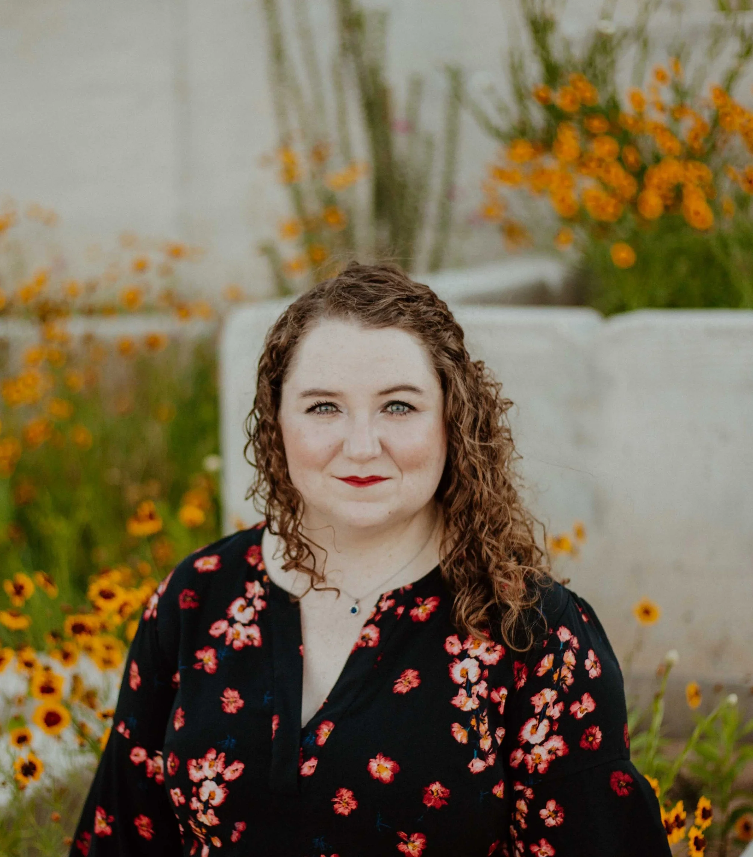 A woman with curly red hair and blue eyes, wearing a black floral top, sitting outdoors among orange and yellow flowers.