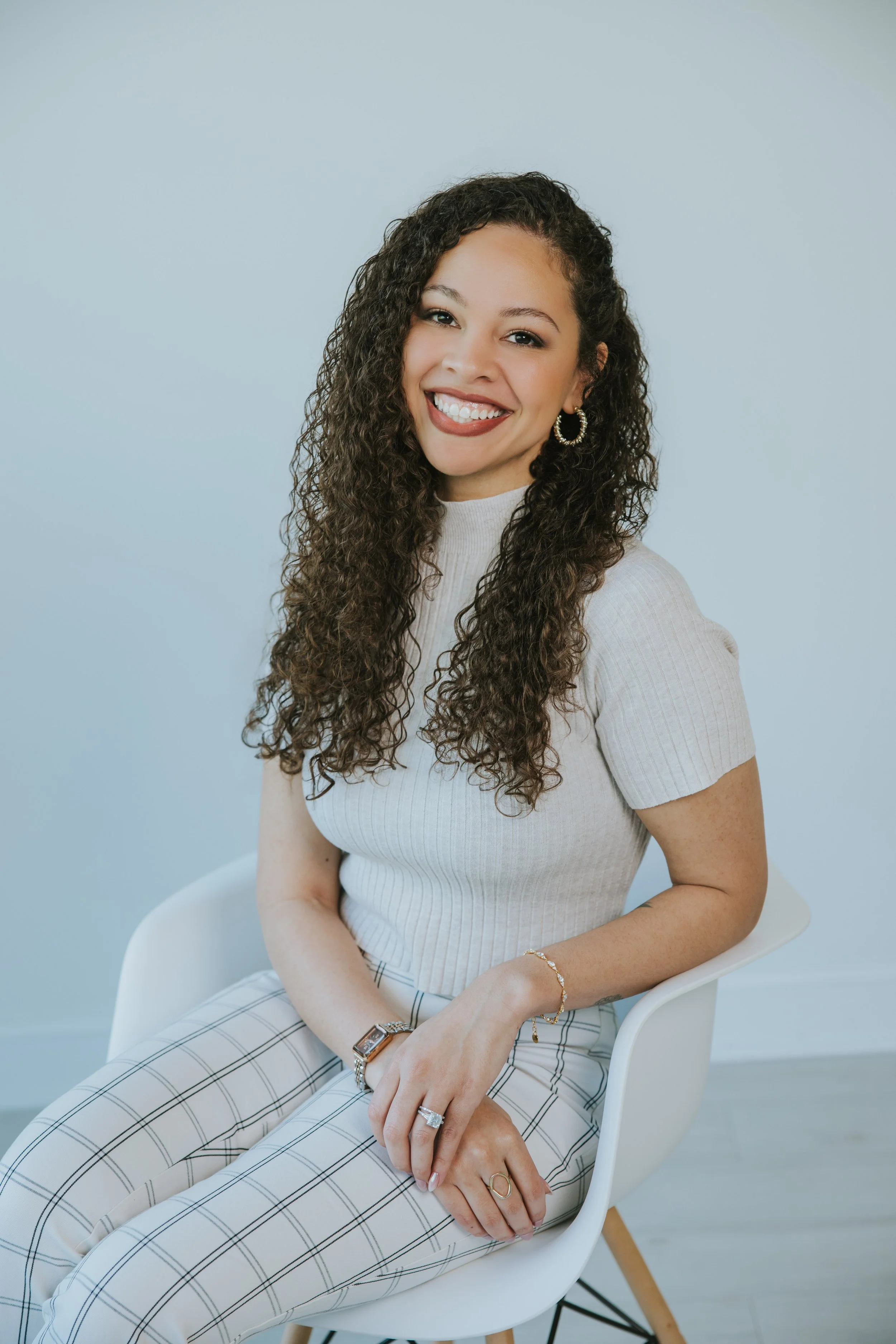 A young woman with curly brown hair and a big smile, wearing a light blue top, sitting on a blue chair in a room with a light gray wall and a small bookshelf with a plant in the background.