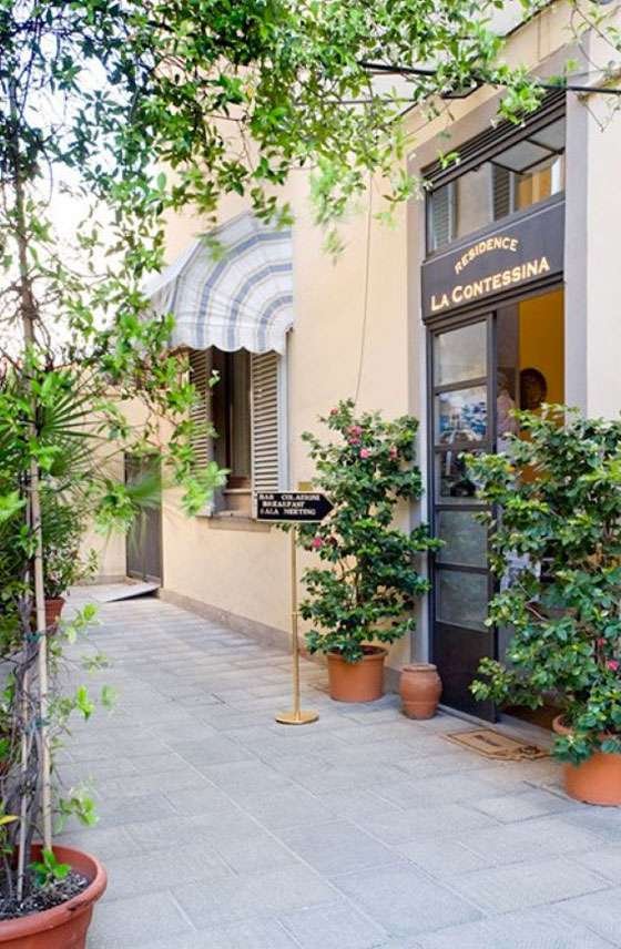 Entrance of Residence La Contessina with potted plants and a signpost, outdoor patio with greenery and striped awning window.