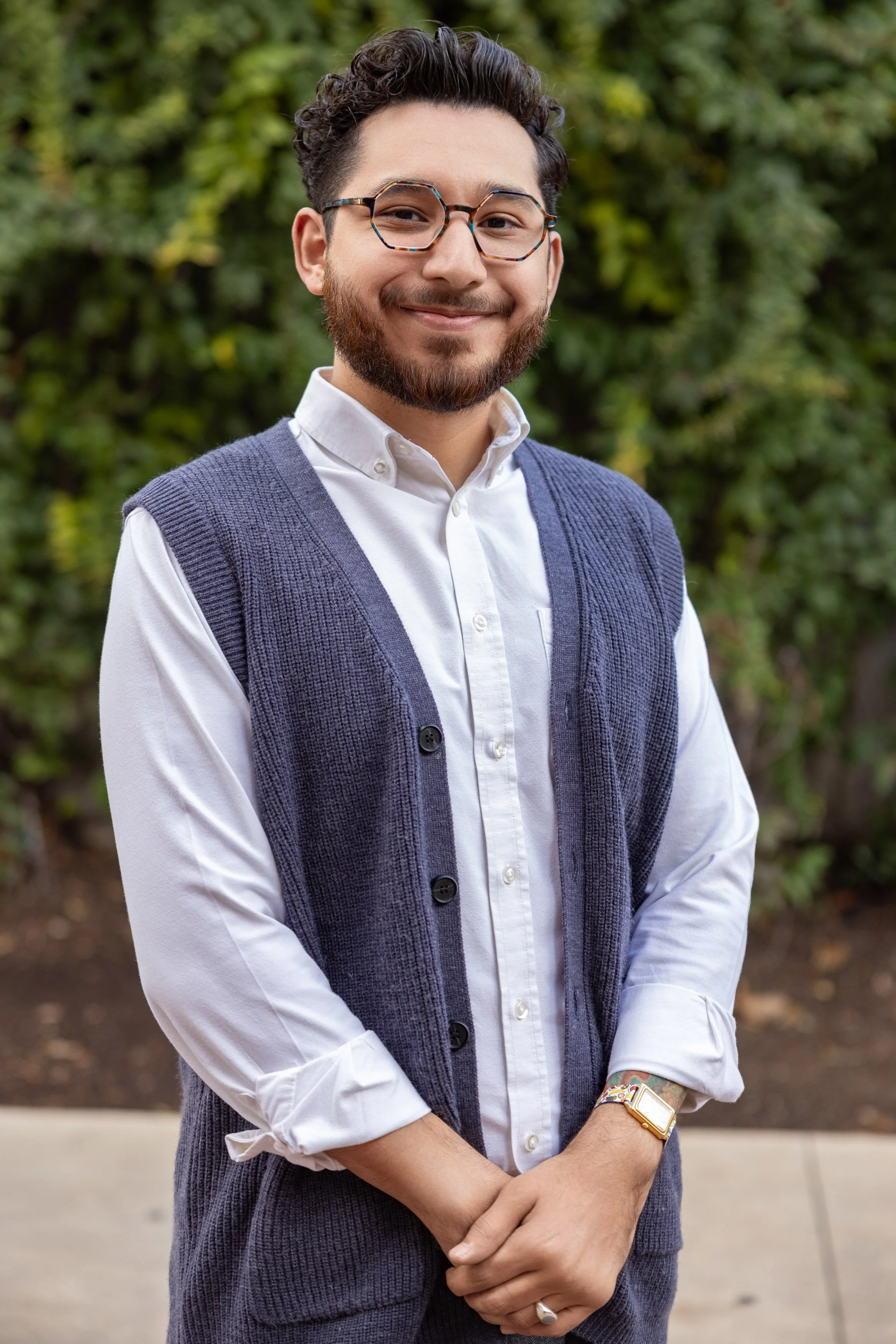 Smiling man with glasses, white shirt, and blue cardigan, standing outdoors with trees in the background.