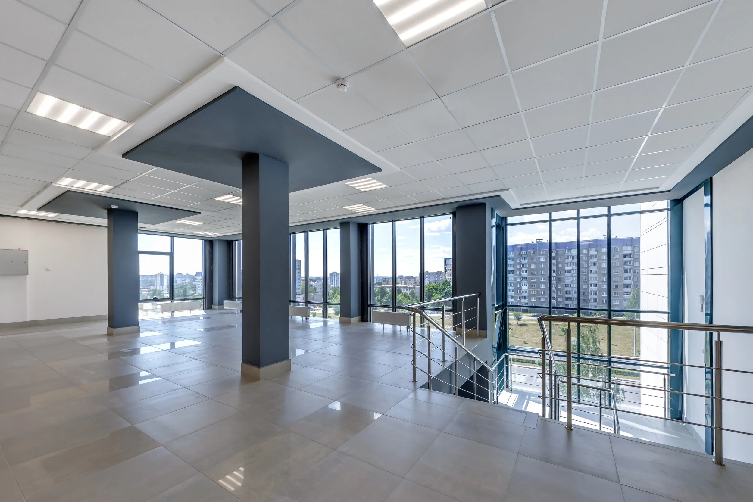Empty modern office space with large windows, tiled floor, white ceiling with lights, blue accent walls, and a staircase with metal railing.