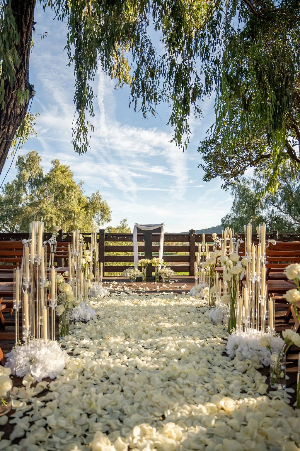 Outdoor wedding ceremony setup with a pathway decorated with white flowers and petals, flanked by tall vases with white flowers and candles, under large trees with a blue sky in the background.