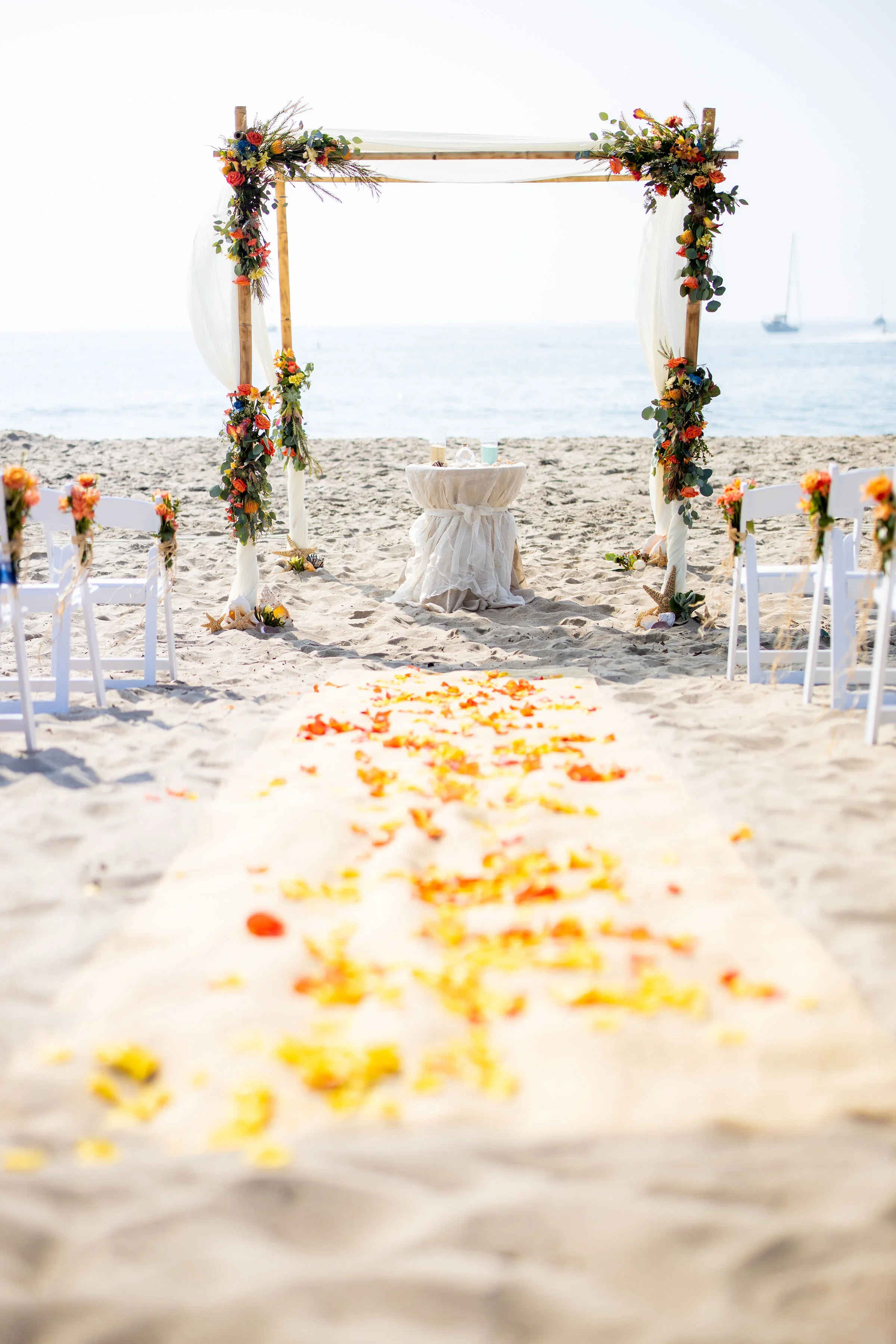 Beach wedding altar decorated with flowers, situated on sandy beach with ocean in the background and sailboats in the distance.