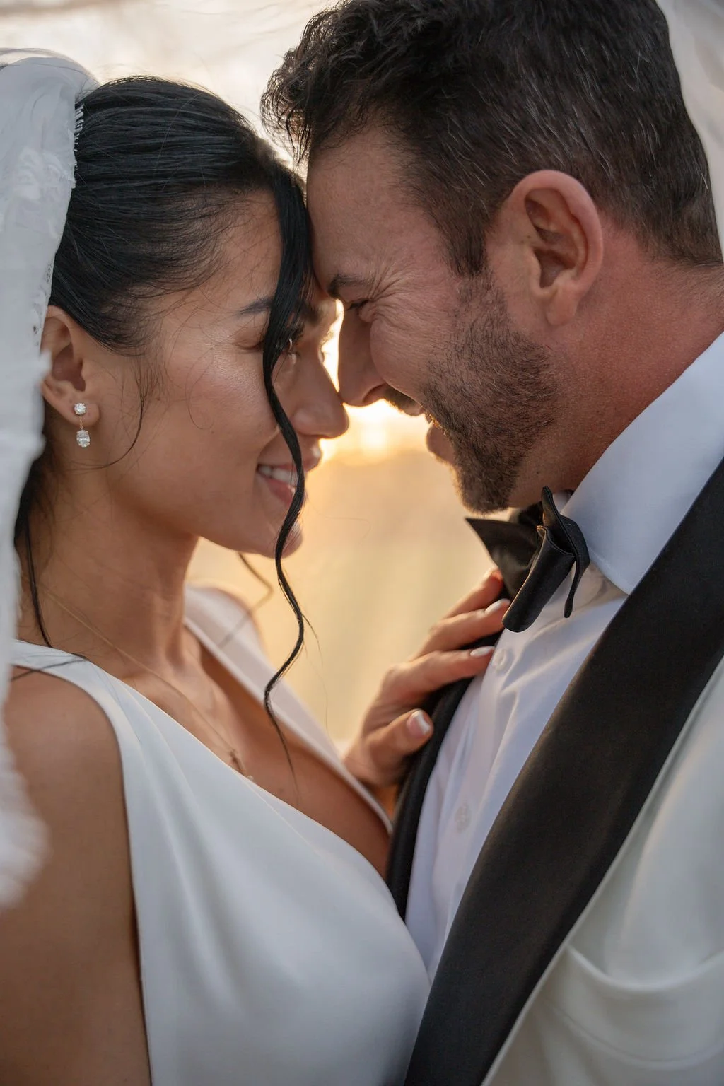 A bride and groom with their foreheads touching, smiling at each other during their wedding, with warm lighting in the background.