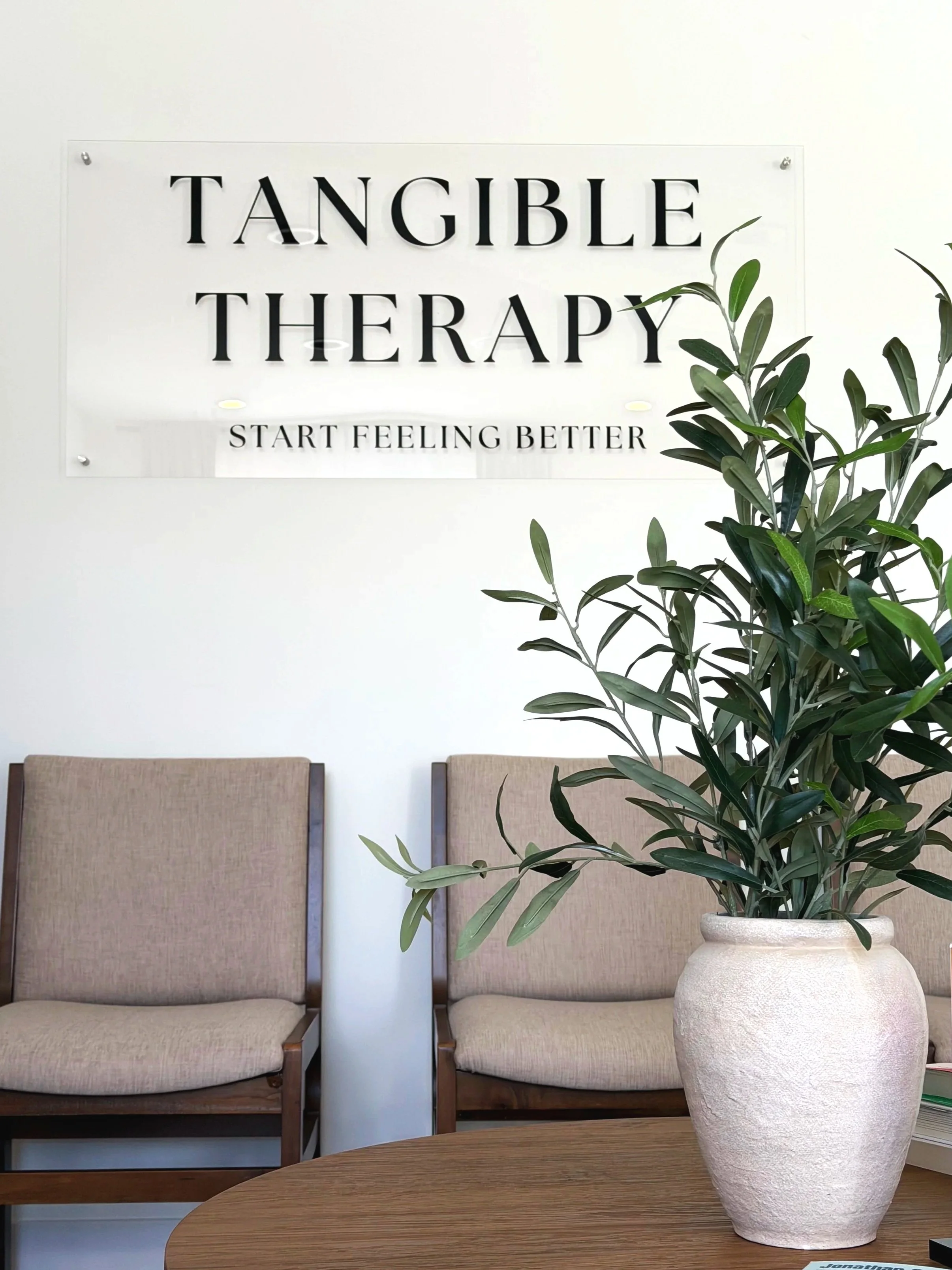 Tangible Therapy waiting room with a potted plant on a oak table with waiting room chairs in the background. All Below a sign on the wall that reads "Tangible Therapy Start Feeling Better"