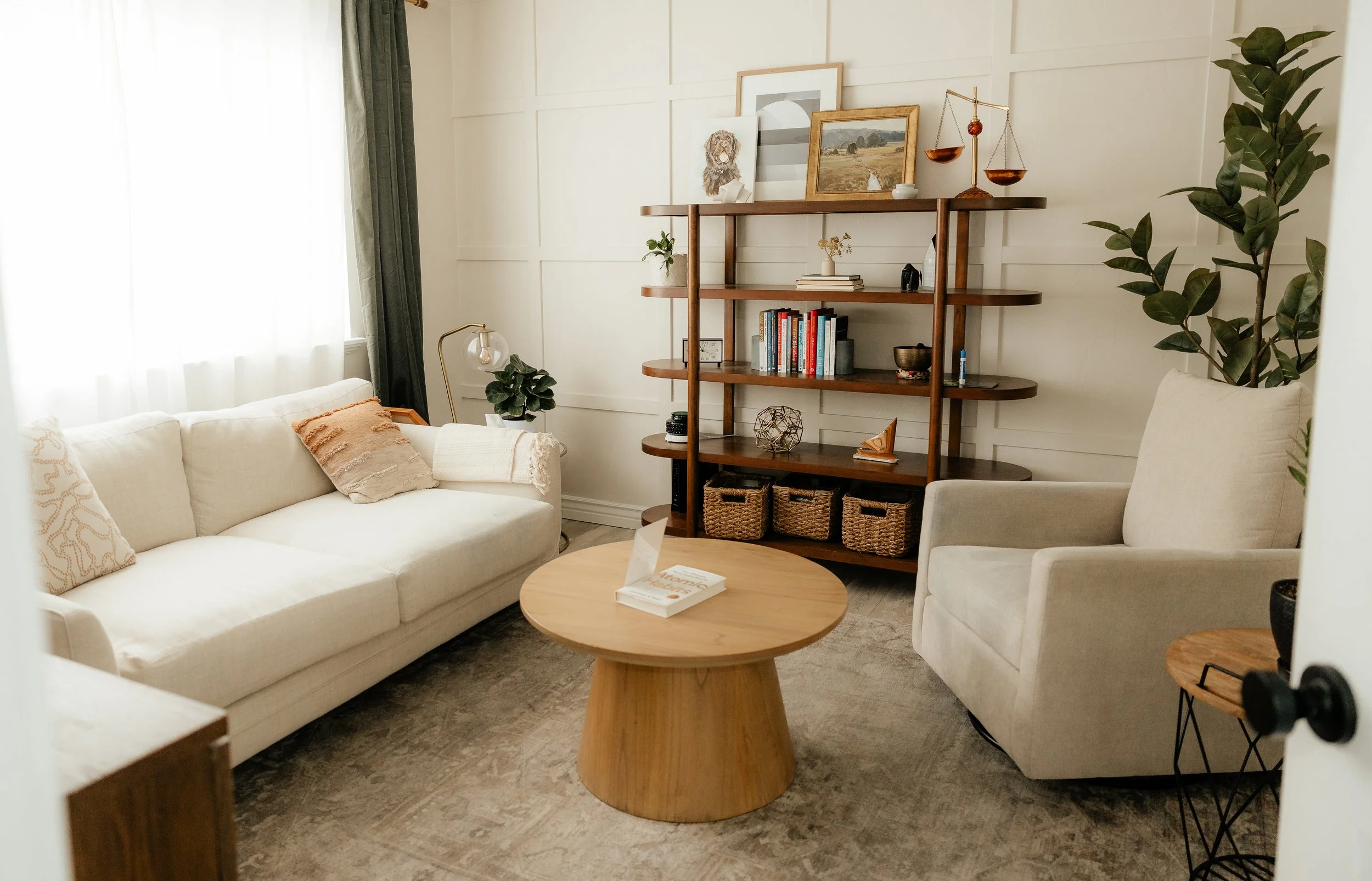 A cozy, minimalist therapy office at Tangible Therapy in Orem Utah features a white couch with cushions, a round wooden coffee table, and a shelf with books, plants, and decorative items.