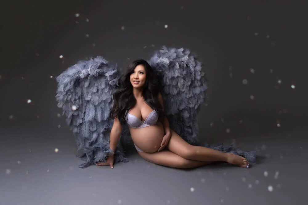 Pregnant woman with long dark hair, wearing lingerie, sitting on the floor, with large gray feathered angel wings against a dark background with glitter during glamorous maternity and pregnancy photography studio session in Columbus ohio