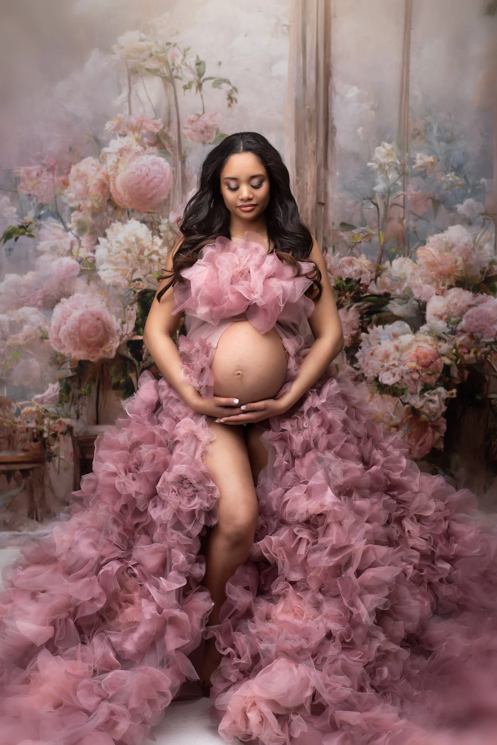 Photo of A pregnant woman with long dark hair and light skin, wearing a pink ruffled dress, holding her belly with both hands, standing amidst a floral backdrop of pink peonies and roses in columbus ohio