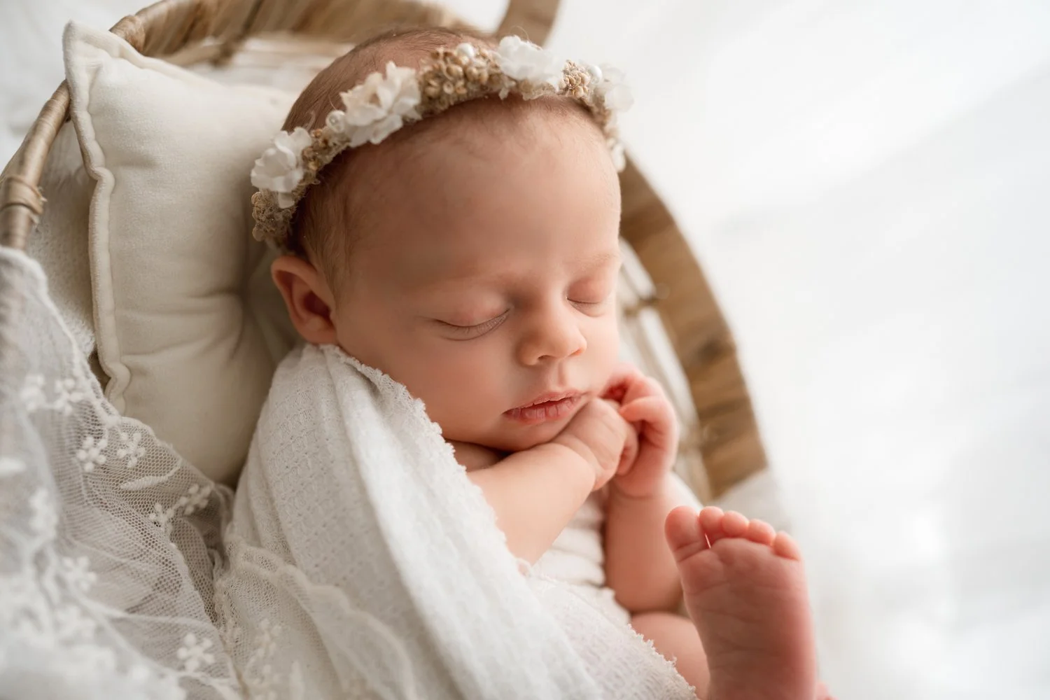 A sleeping baby with a floral headband, resting on a cream-colored pillow, wrapped in a white blanket, in a cozy setting during newborn photography session in Columbus ohio