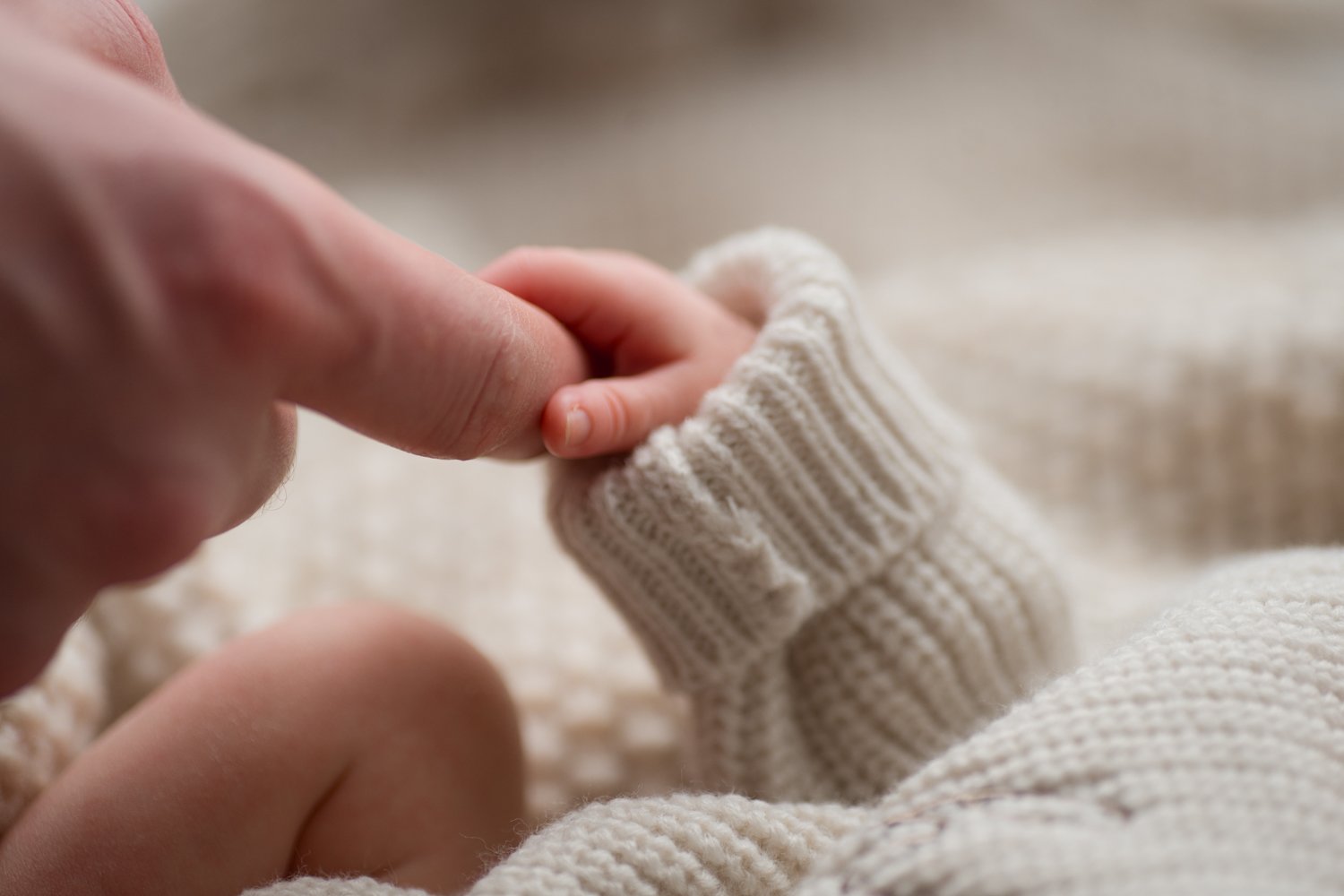 A close-up of an adult holding a baby's tiny hand, both wearing cozy, knitted cream-colored clothing during lifestyle session at Columbus ohio newborn photography studio Wildflower Fine Art Portraits