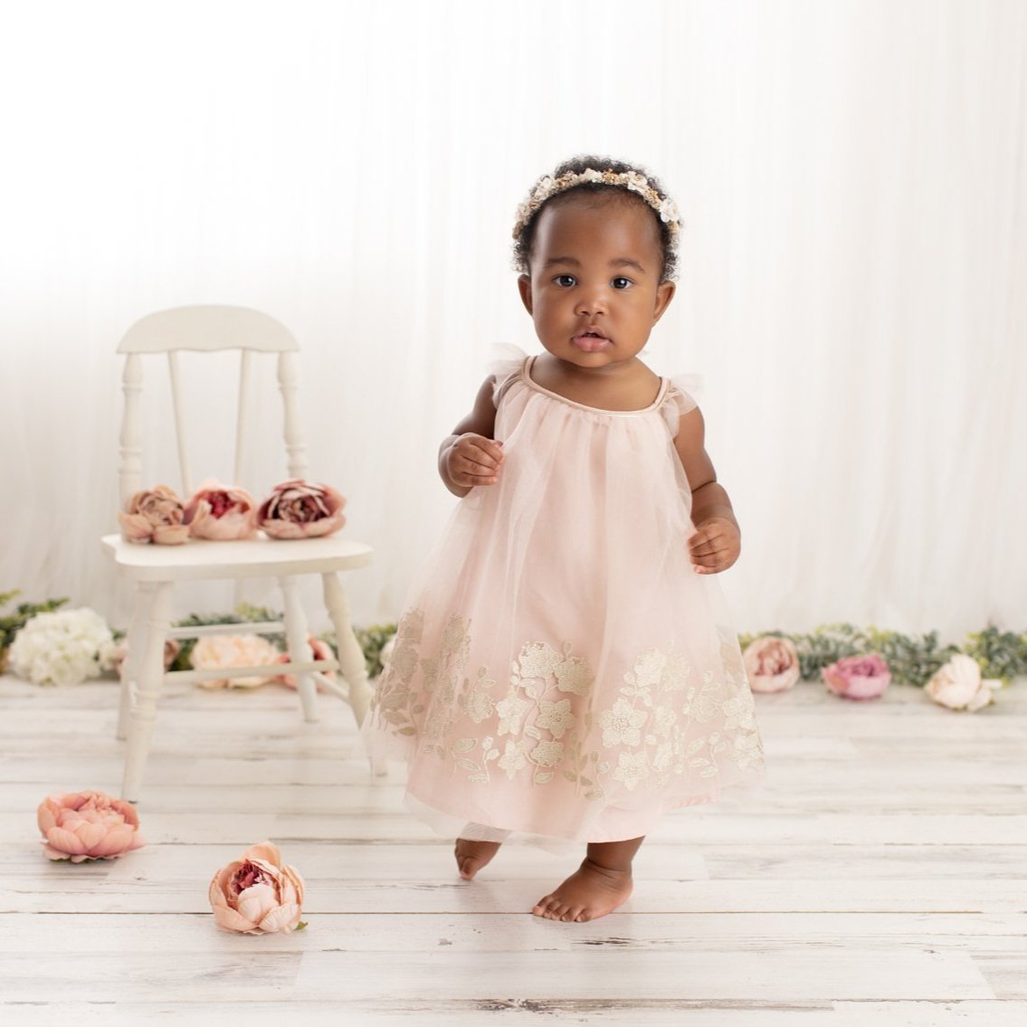 A young girl in a light pink dress with floral embroidery, standing barefoot on a white wooden floor in a studio with a white backdrop and floral decorations.