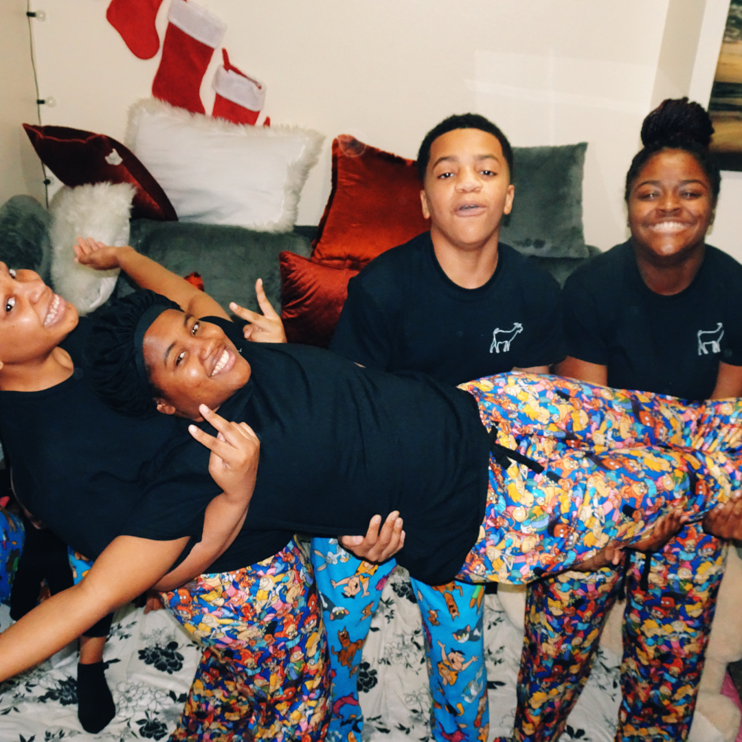 Four people, three women and one man, are sitting on a couch in festive pajamas. One woman is lying across the laps of the others, smiling and making peace signs. The background features Christmas stockings and pillows.