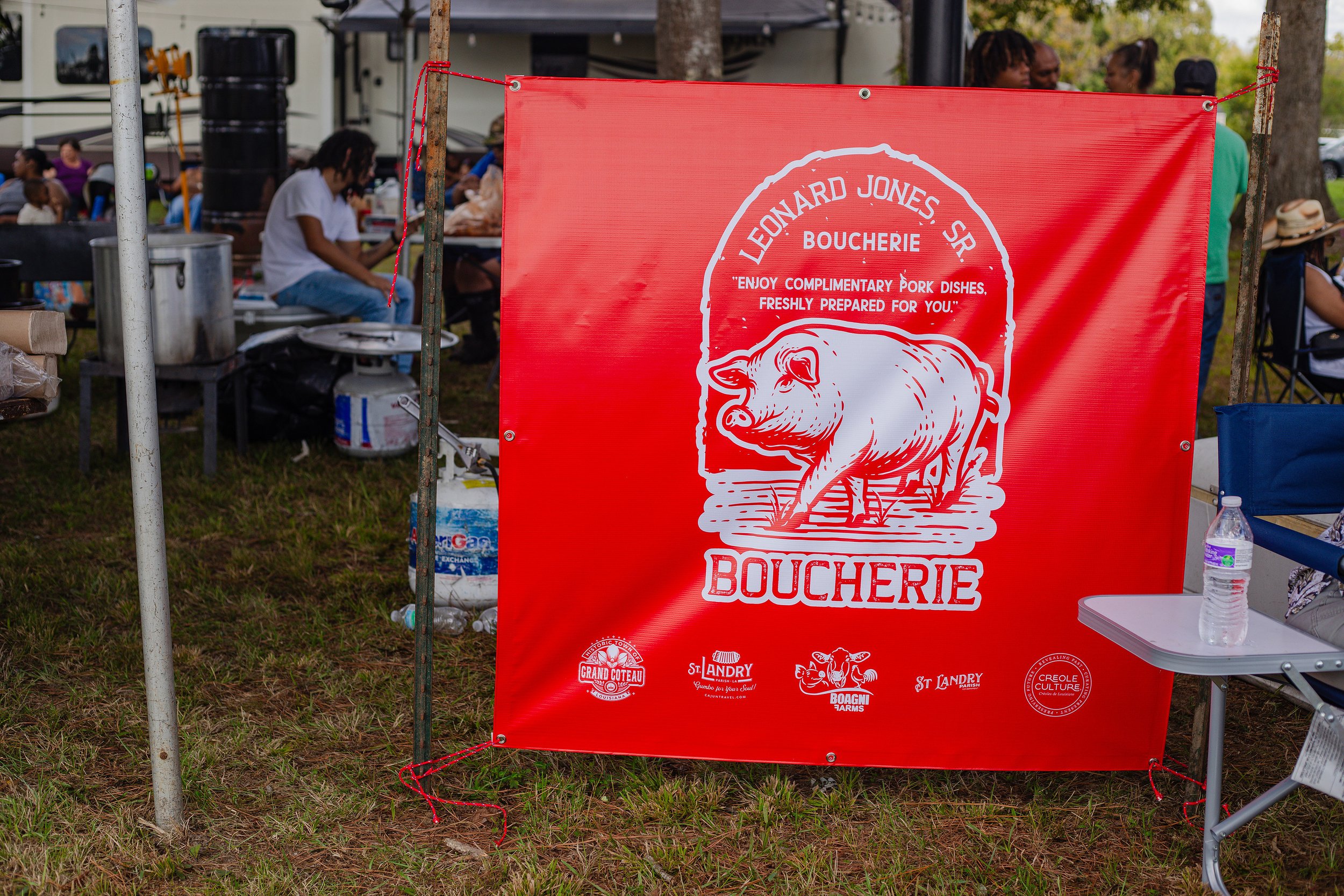Red banner with white graphic of a pig, advertising Leonard Jones Sr. Boucherie, a butcher shop selling pork dishes. The banner is outdoors at a gathering with people and tents in the background, and has logos at the bottom for Grand Coteau, St. Andr