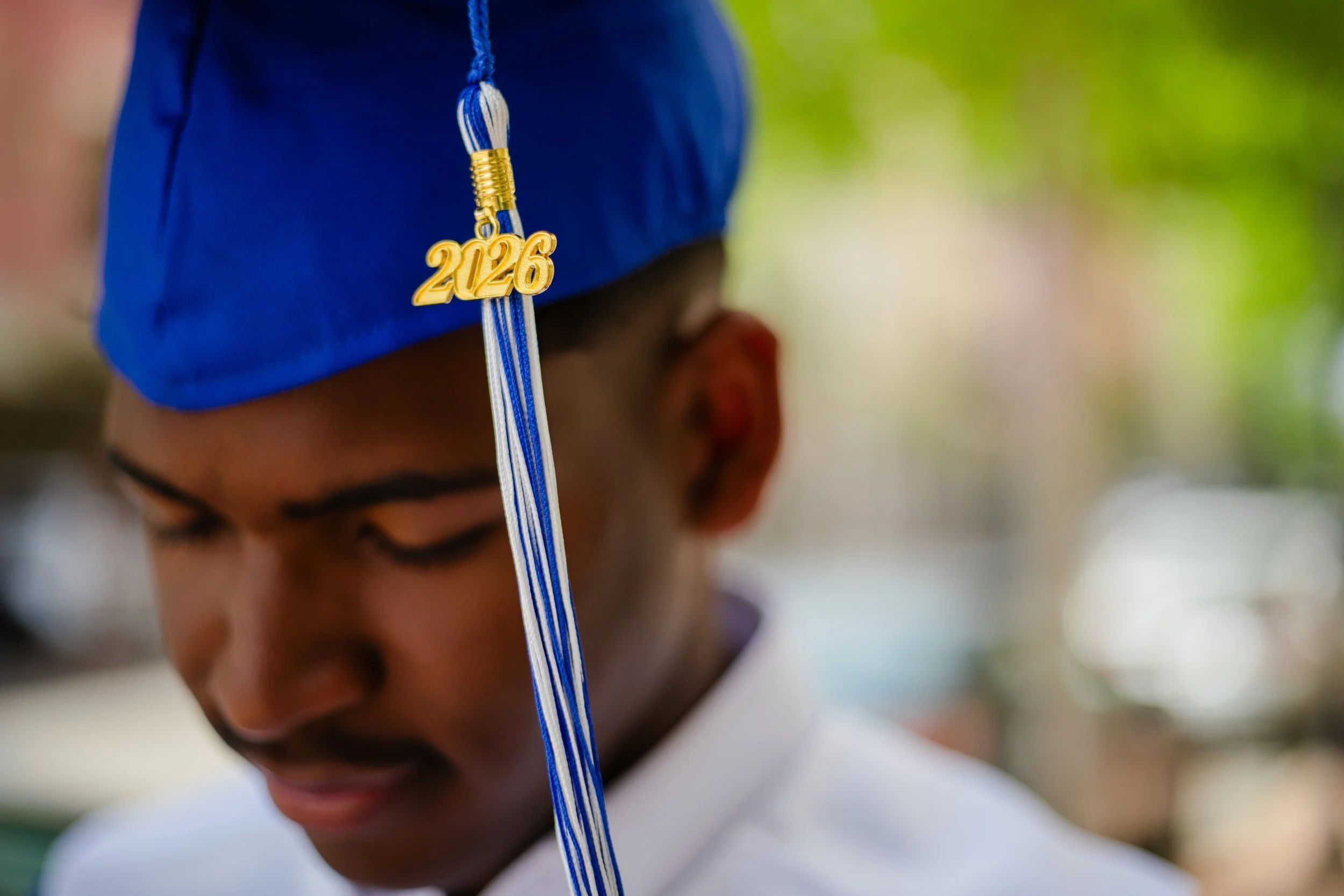 Graduate wearing a blue cap with a gold and blue tassel featuring the year 2026, outdoors with blurred background.
