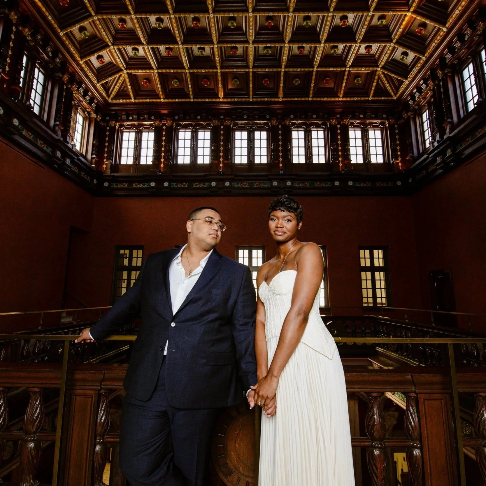 A man in a dark suit and a woman in a white strapless dress holding hands inside a grand, historic building with intricate woodwork and large windows.