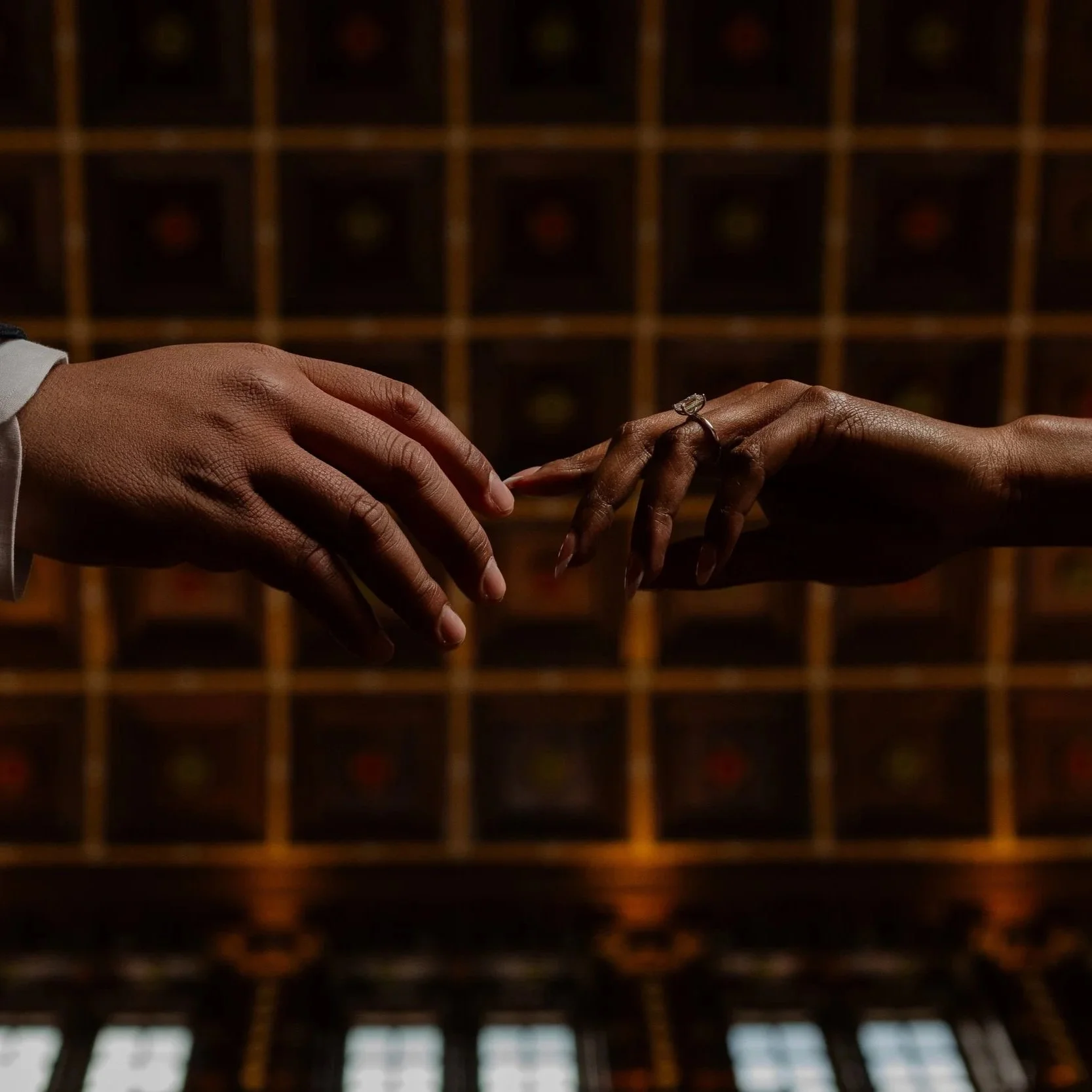 Two hands reaching towards each other, one with a wedding ring, in front of a dark wooden background with small compartments.