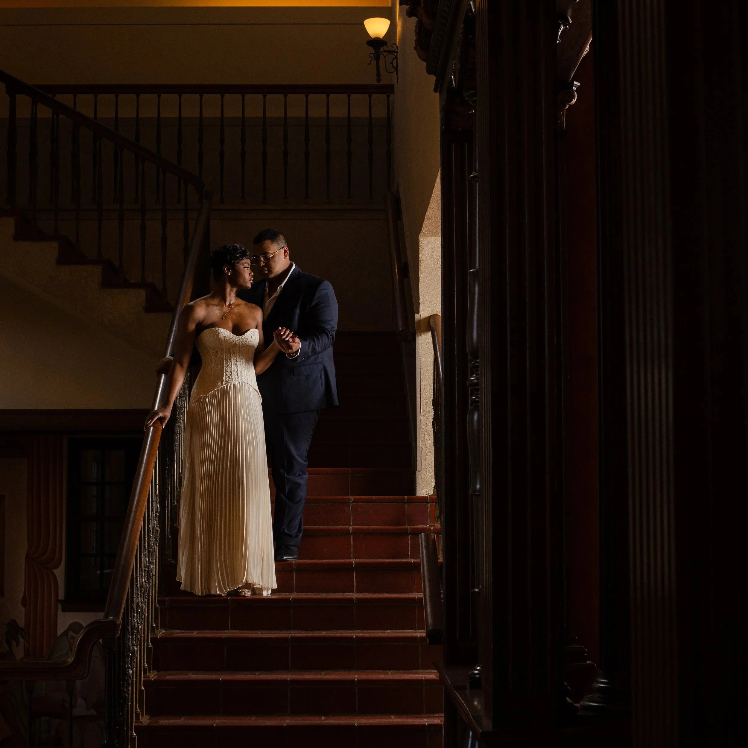 A couple standing on a staircase in an elegant dimly lit interior, holding hands, with the woman wearing a strapless cream gown and the man in a dark suit.