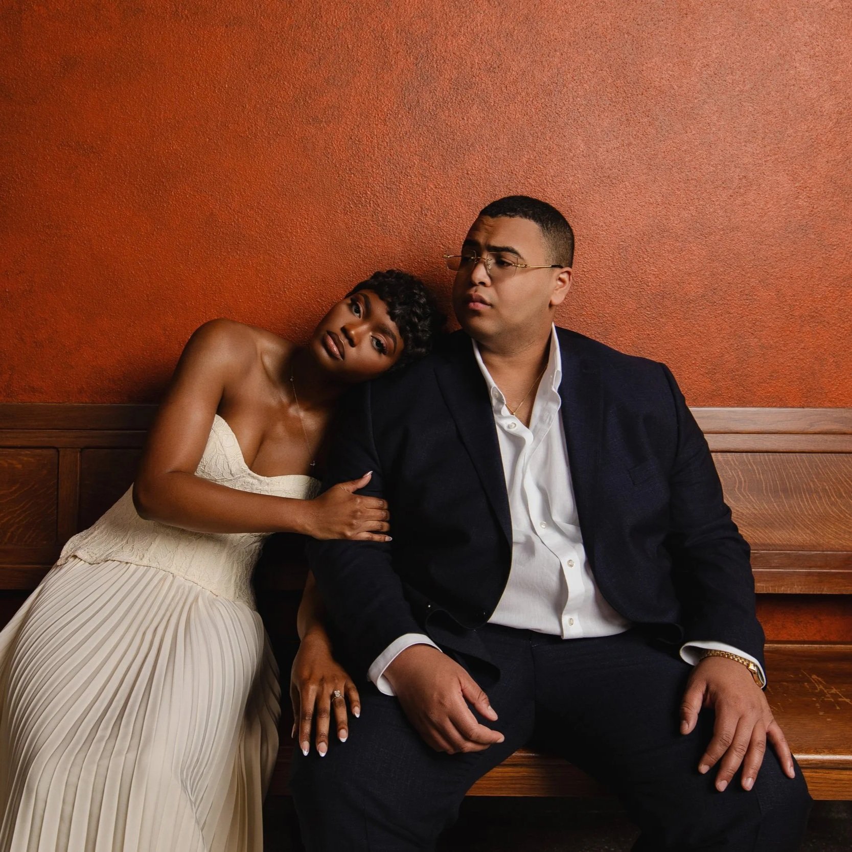 A woman in a strapless beige dress leaning on a man in a black suit and white shirt, sitting on a wooden bench against a textured orange wall.
