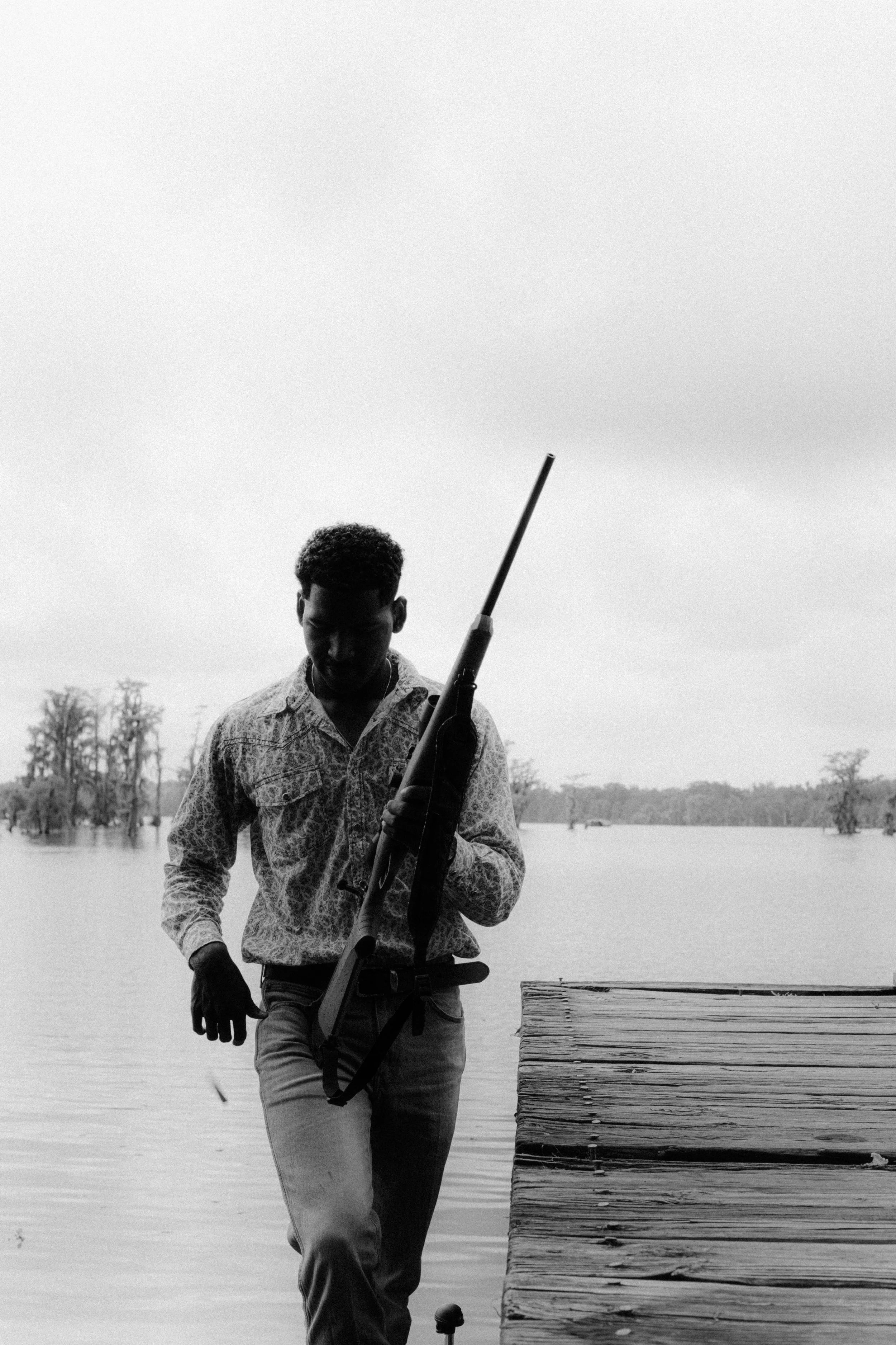 A man holding a rifle standing on a wooden dock by a large body of water, with trees in the background, black and white photograph.
