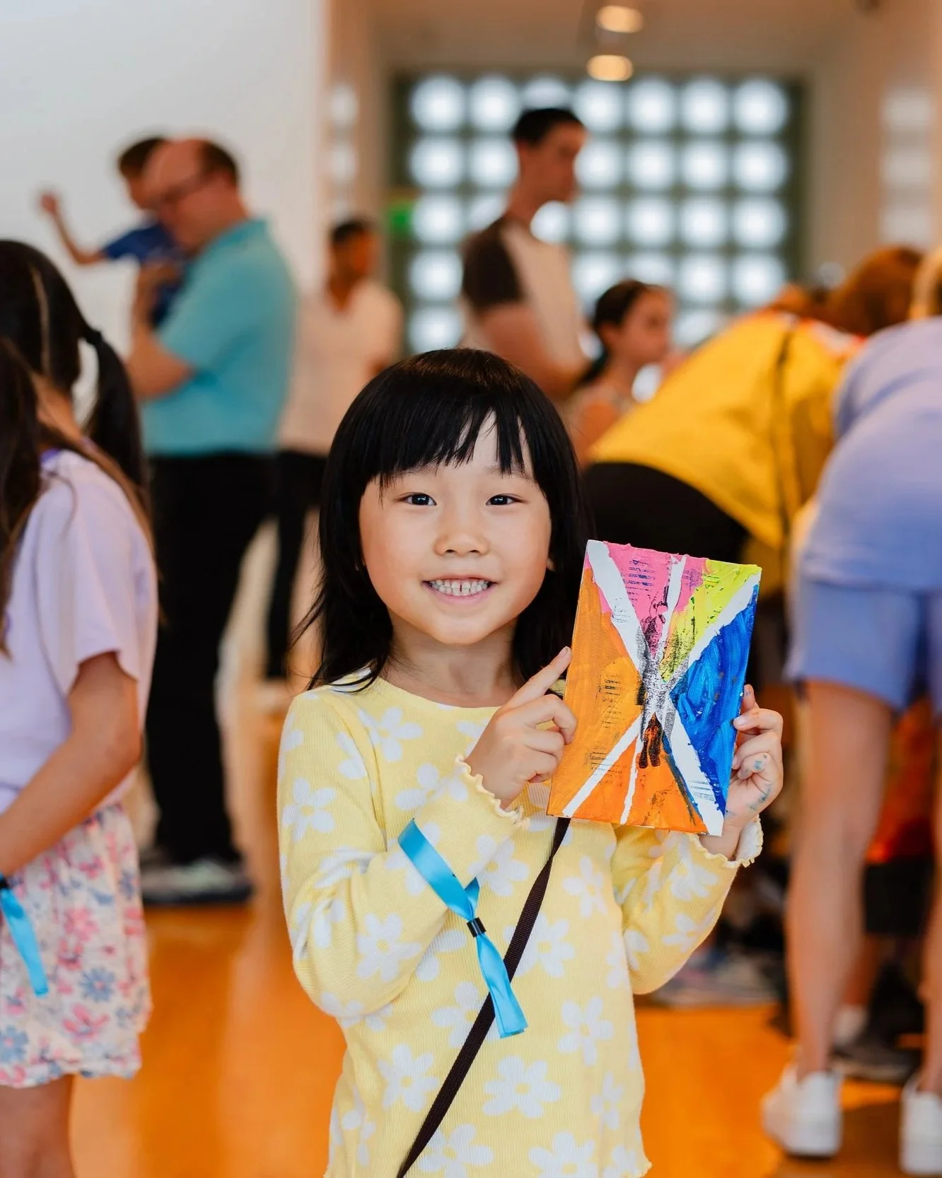 Young girl holding a colorful painted picture, smiling at the camera in a crowded indoor setting.