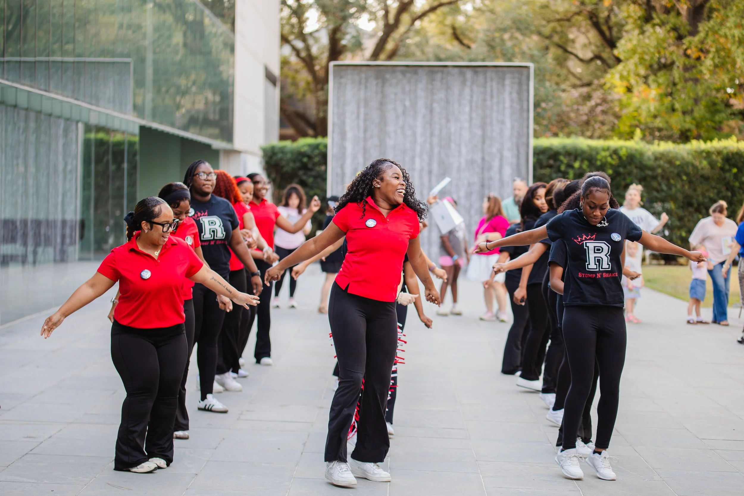 Group of women dancing outdoors on a paved area, with trees and a modern building in the background.