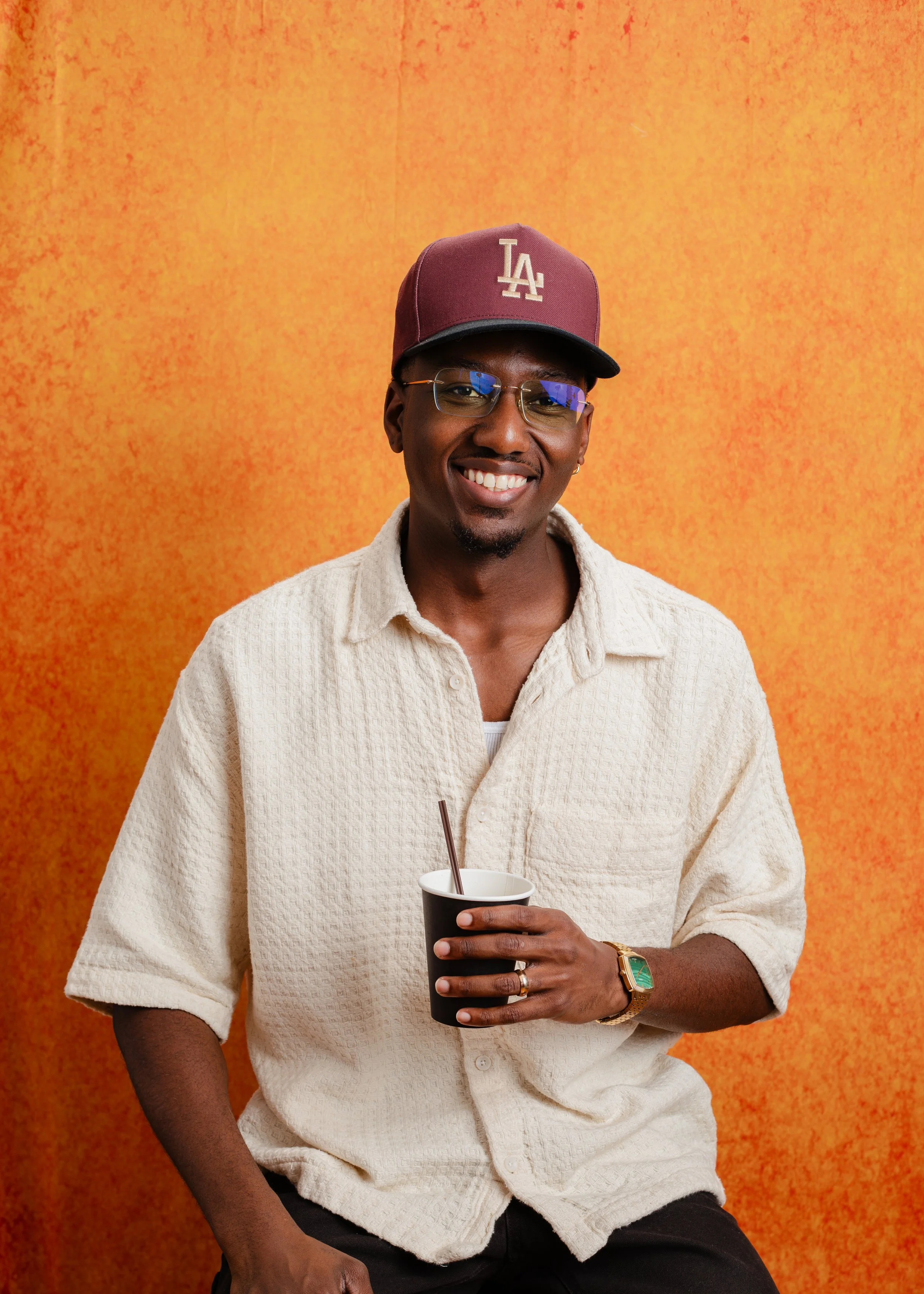A young man smiling and holding a cup with a straw, standing against an orange background. He's wearing glasses, a maroon Los Angeles Dodgers cap, a beige textured shirt, a gold watch, and a ring.
