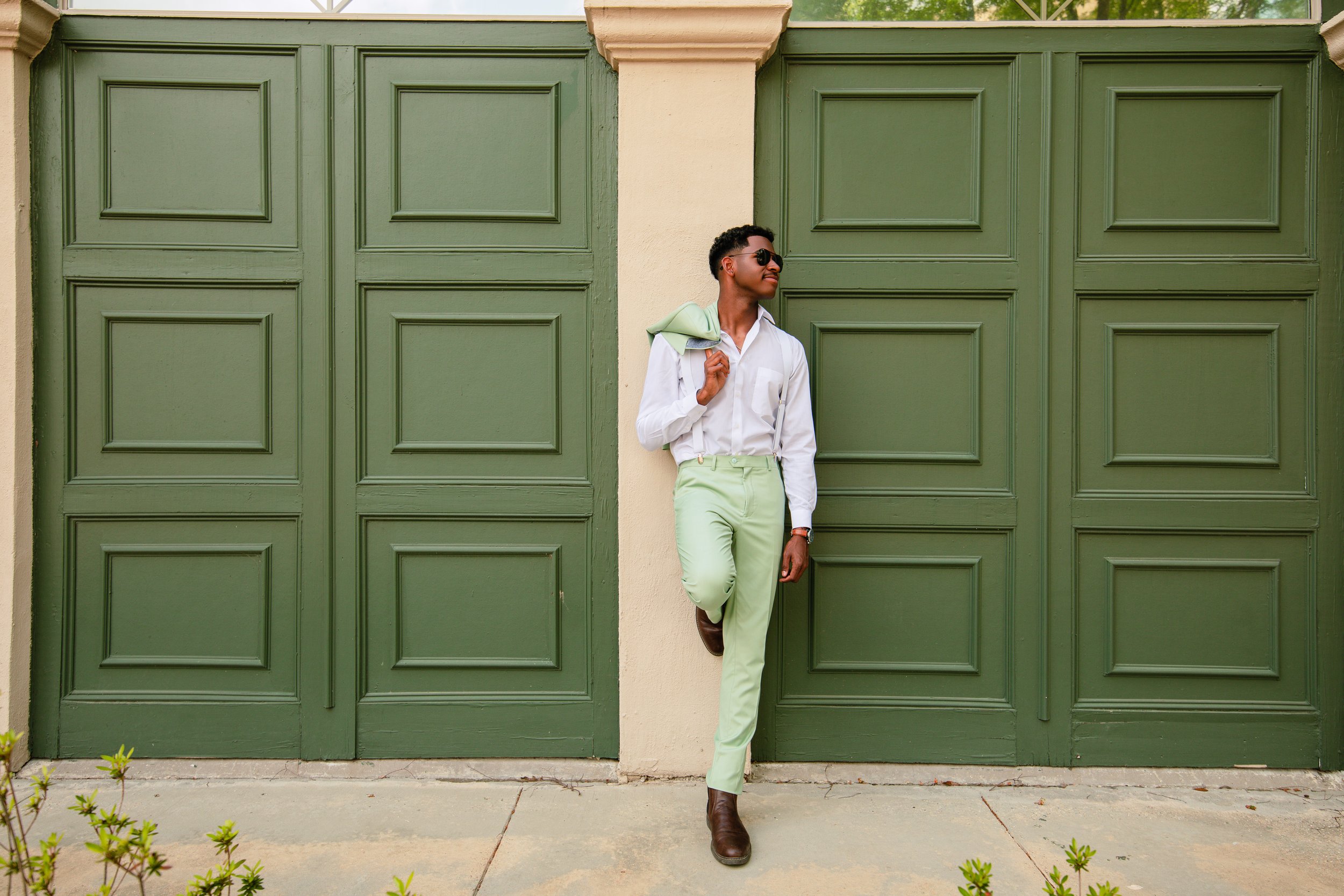A young man standing in front of green garage doors, wearing light green pants, a white shirt, brown boots, sunglasses, and holding a green jacket over his shoulder.