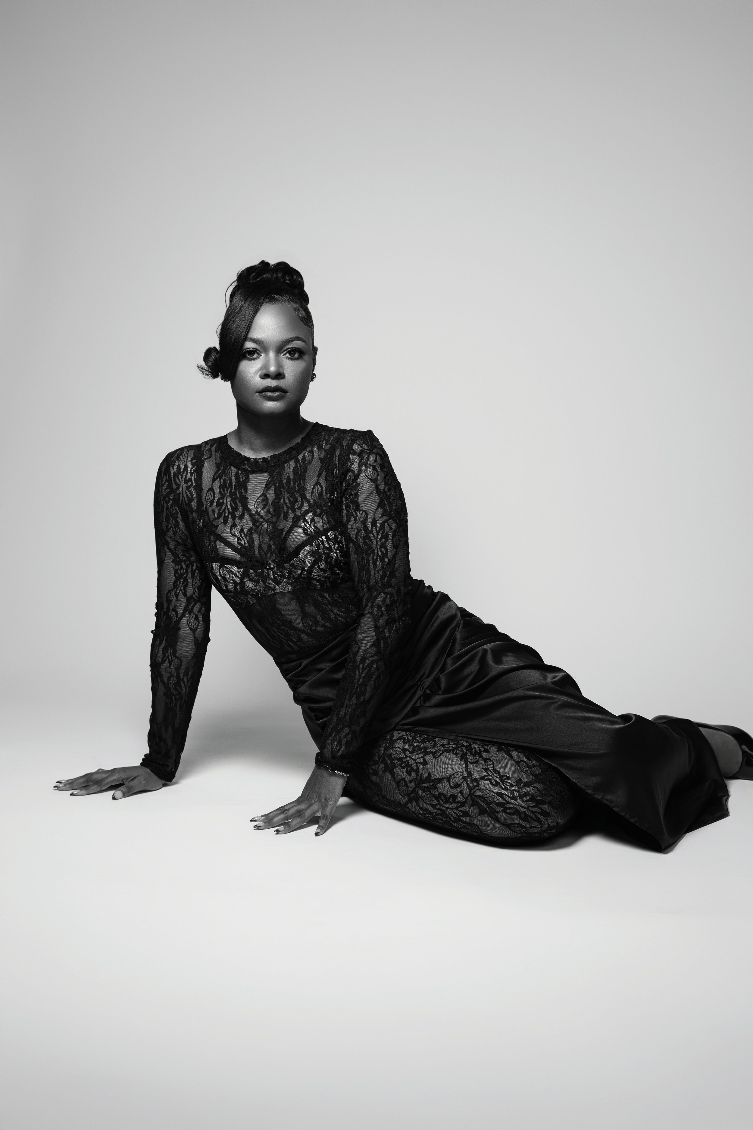 Black and white photo of a woman in a lace top and satin skirt, seated on the floor, with one hand resting on the ground and the other slightly bent, gazing directly into the camera, against a plain gray background.