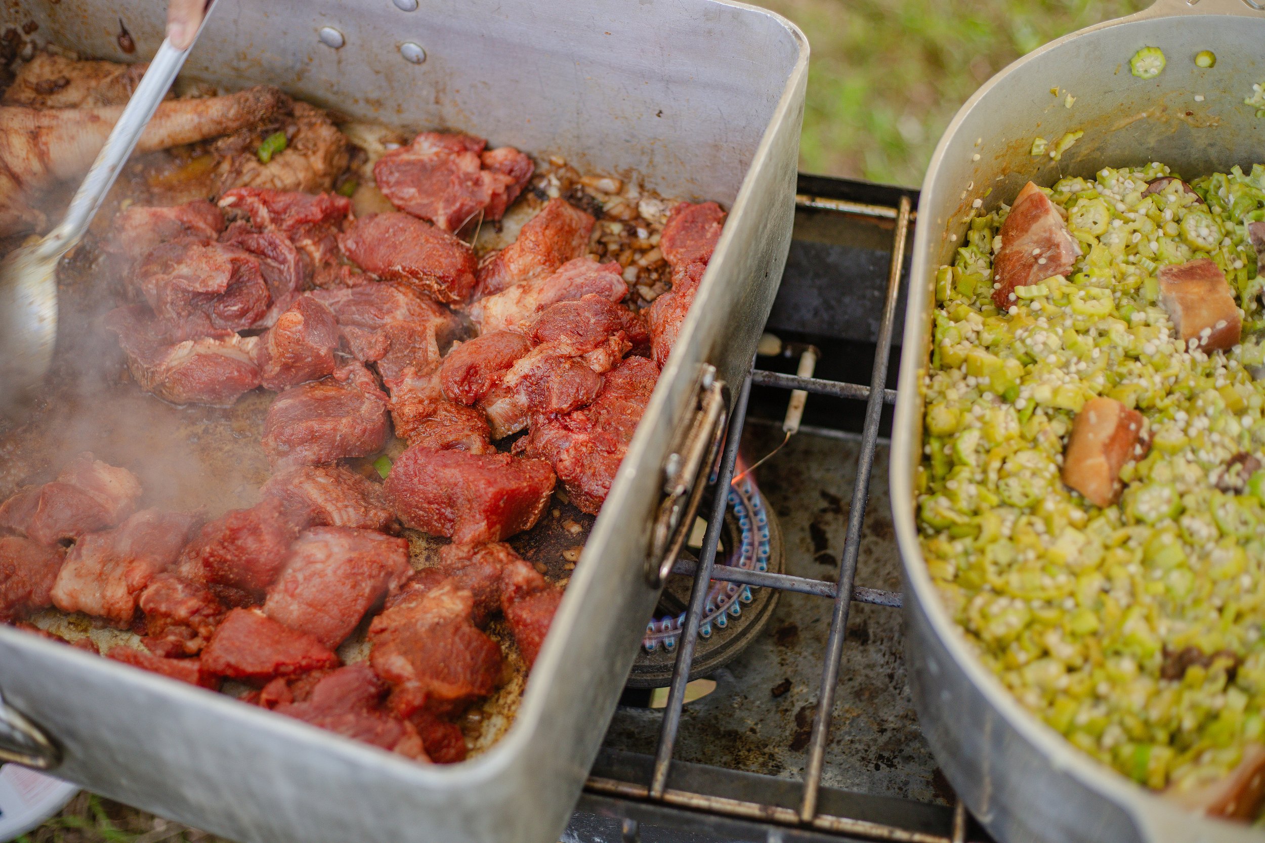 Cooking meat and vegetables outdoors on a grill with two metal trays filled with cooked meat and chopped vegetables.