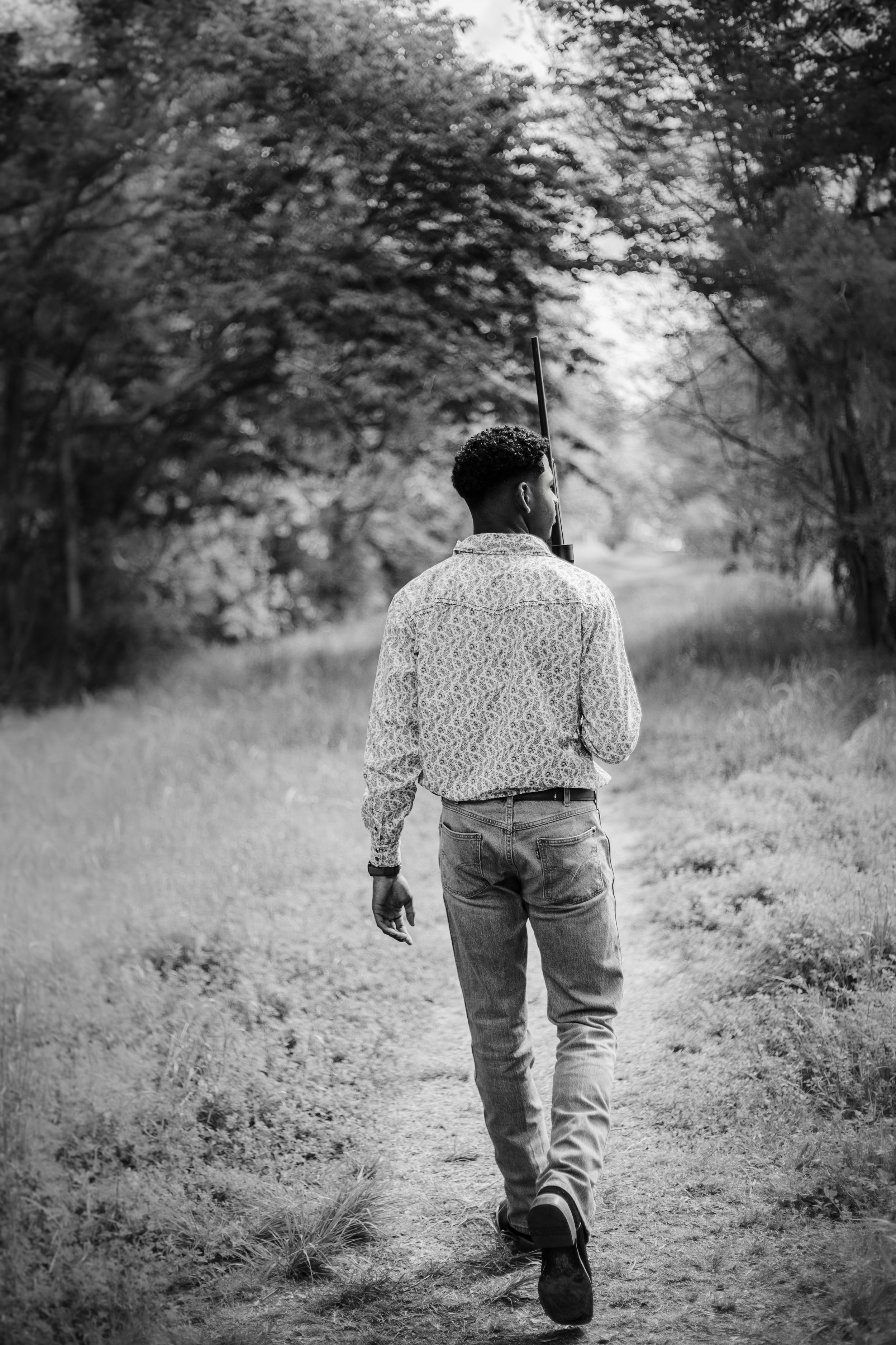 A man walking down a dirt path surrounded by trees, carrying a rifle on his shoulder, in black and white.