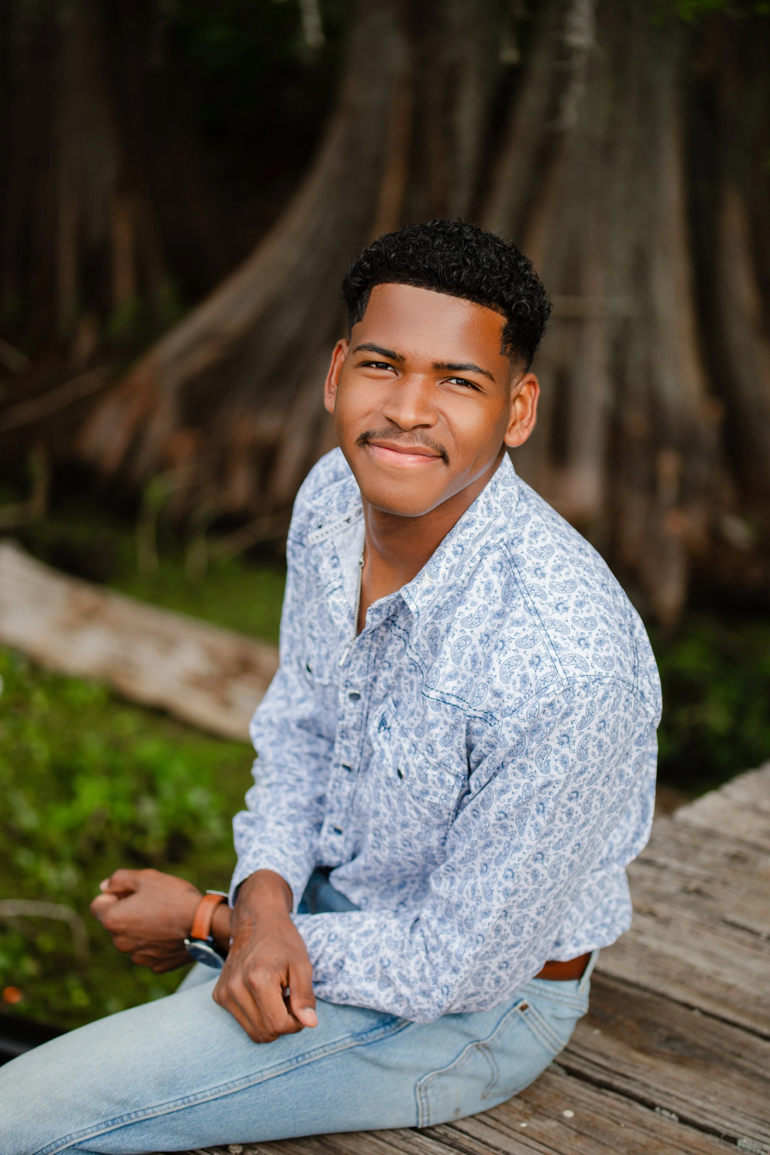 A young man with short curly hair and a mustache, wearing a light blue patterned shirt and light blue jeans, sitting on a wooden dock outdoors in front of large tree roots.