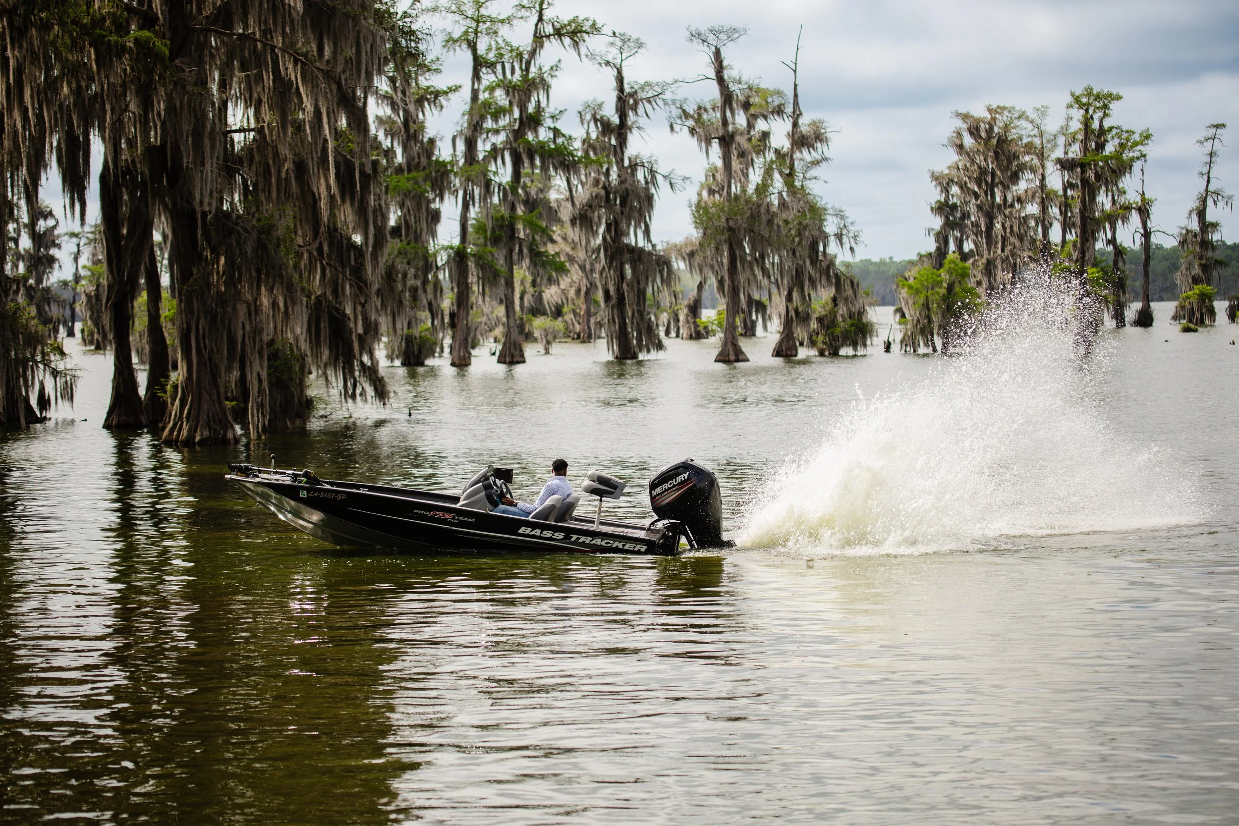 A person driving a black Bass Tracker boat with a Mercury outboard motor on a lake with cypress trees draped in Spanish moss in the background, creating a splash in the water.