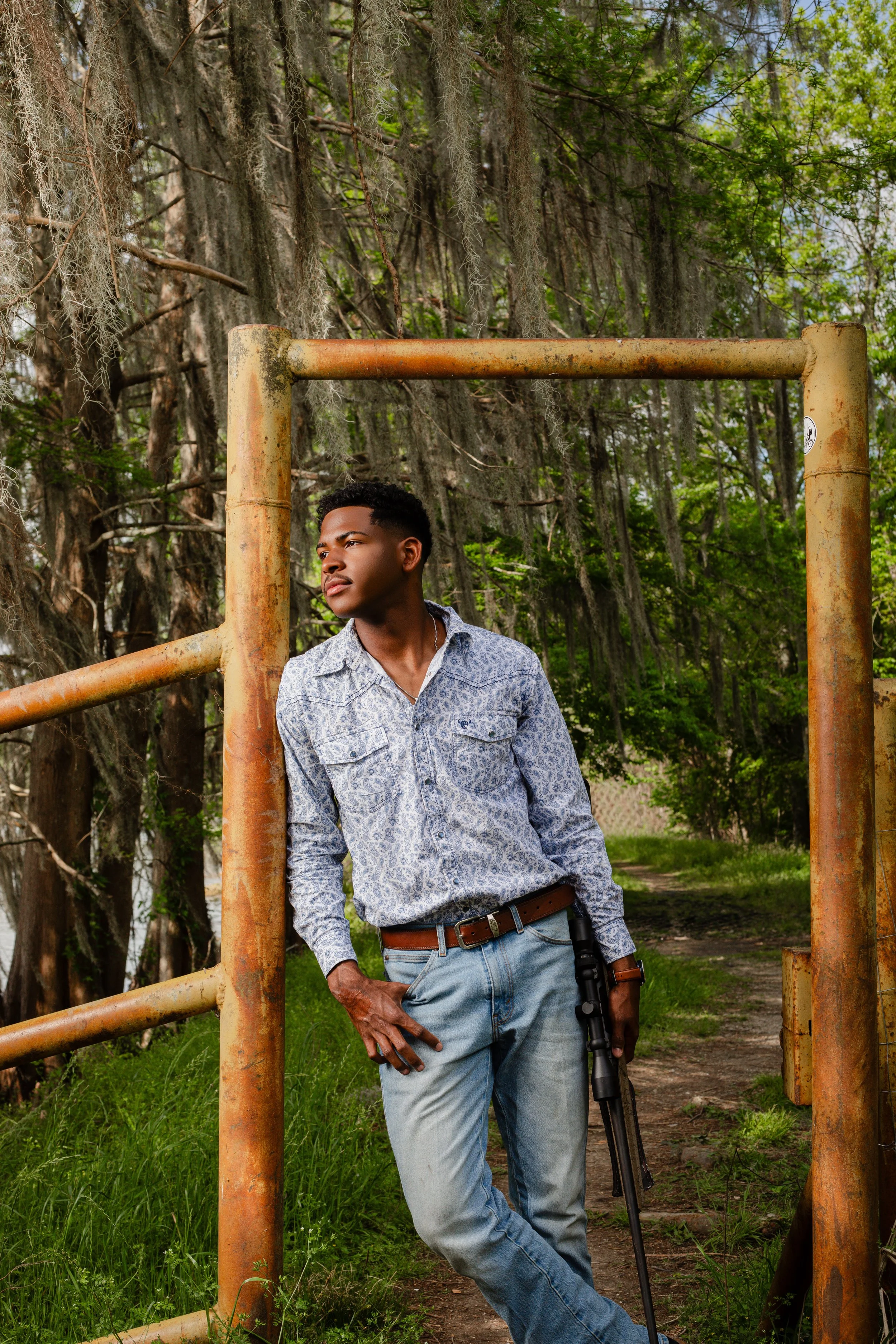 Young man standing outdoors by a rusty gate in a wooded area, holding a rifle in his right hand, looking thoughtfully into the distance.