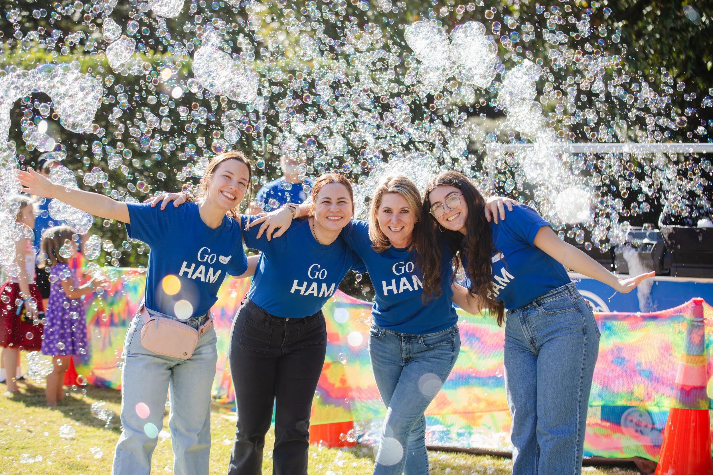 Four women wearing blue shirts with 'Go Ham' printed on them, smiling with arms around each other, standing outdoors at a bubble-blowing event on a sunny day.