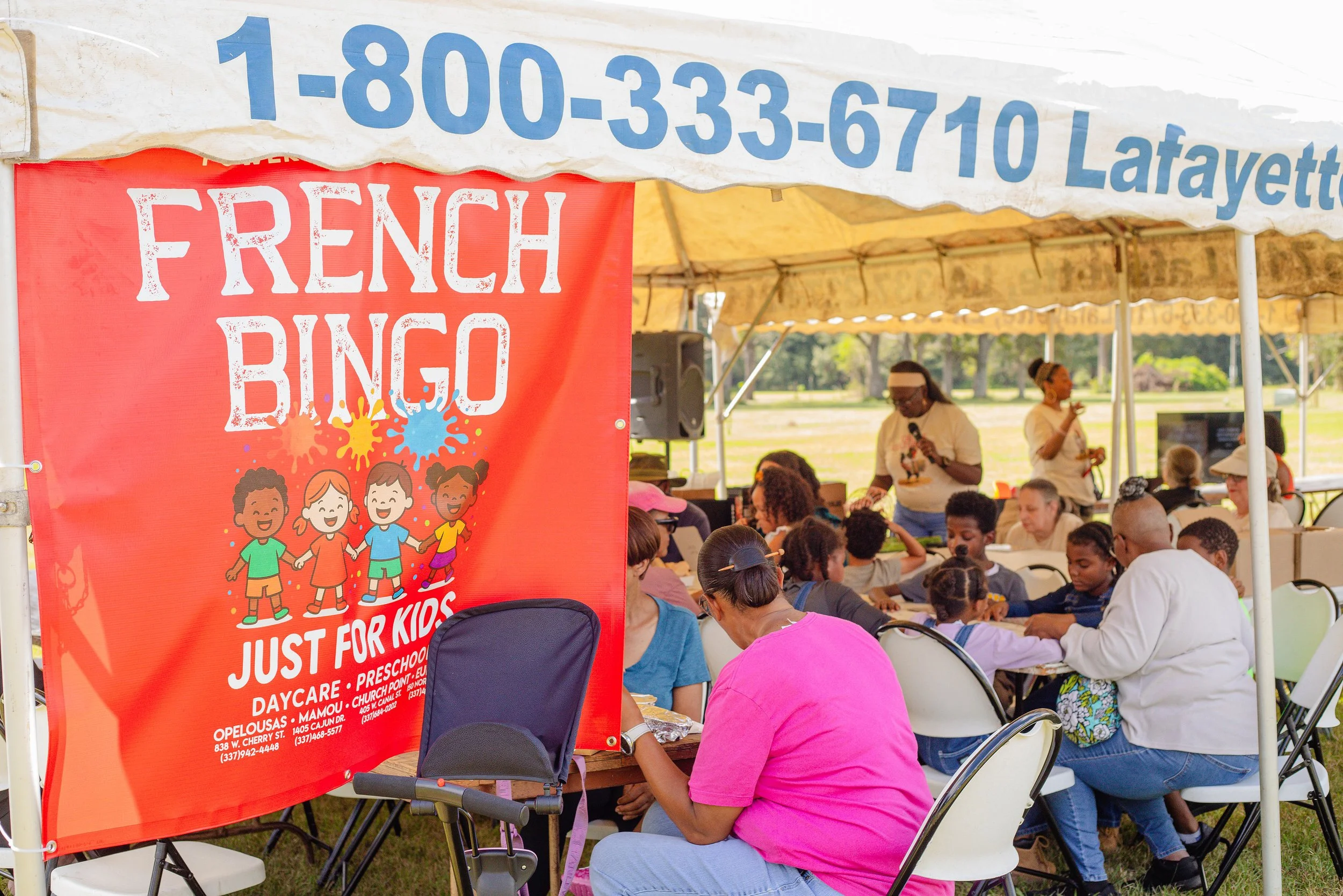 Children and adults playing French Bingo at an outdoor event under a canopy, with a red banner for a daycare. People are sitting at tables, with some standing or speaking into microphones.
