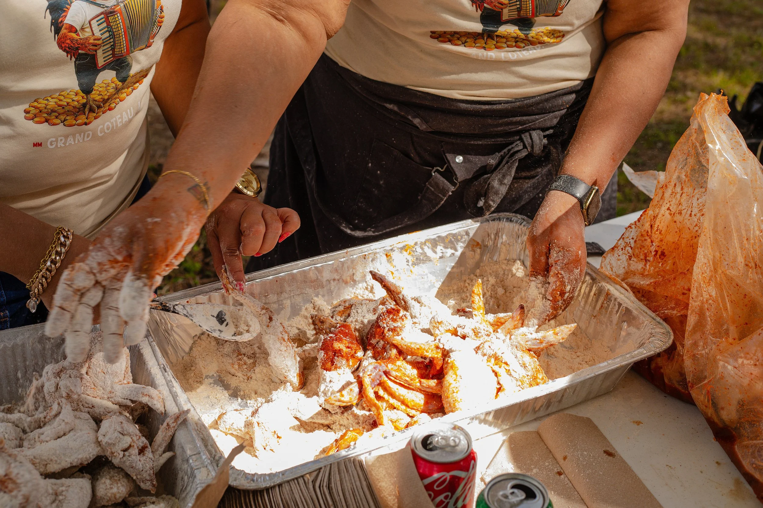Two people preparing fried chicken outdoors, coating chicken pieces with flour in a large aluminum tray, with a soda can and other ingredients on the table.