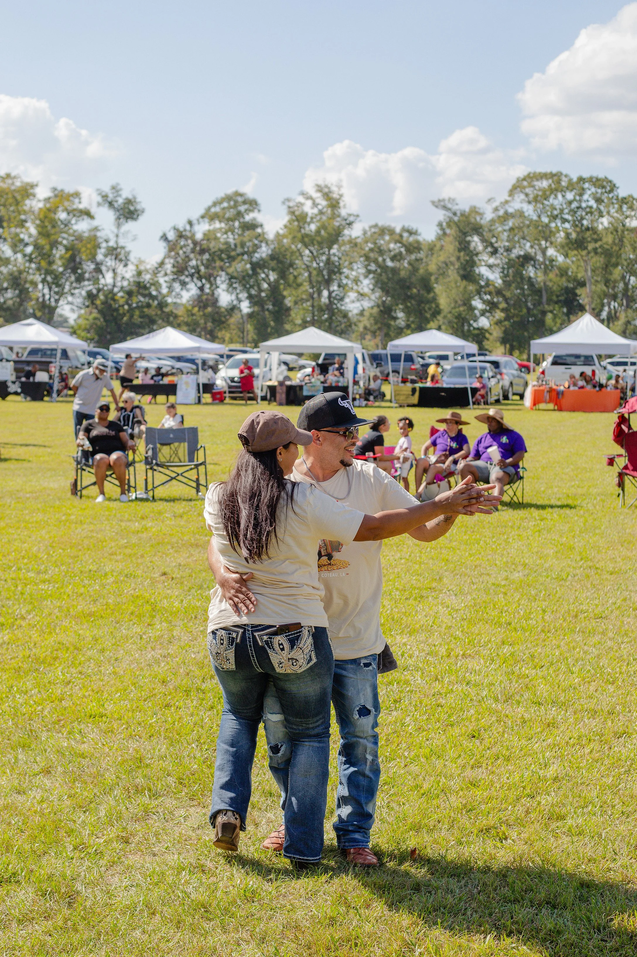 A couple dancing on a grassy field at an outdoor festival, with tents and seated people in the background on a sunny day.
